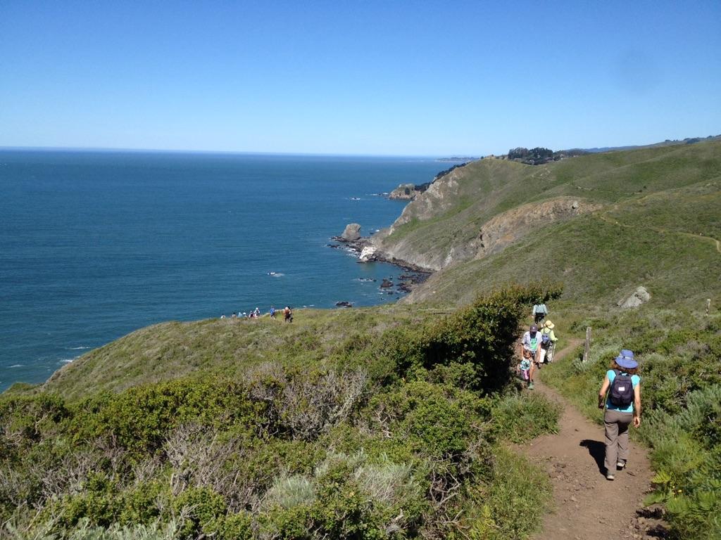 People hike a sunny, green coastal trail above a vast blue ocean.