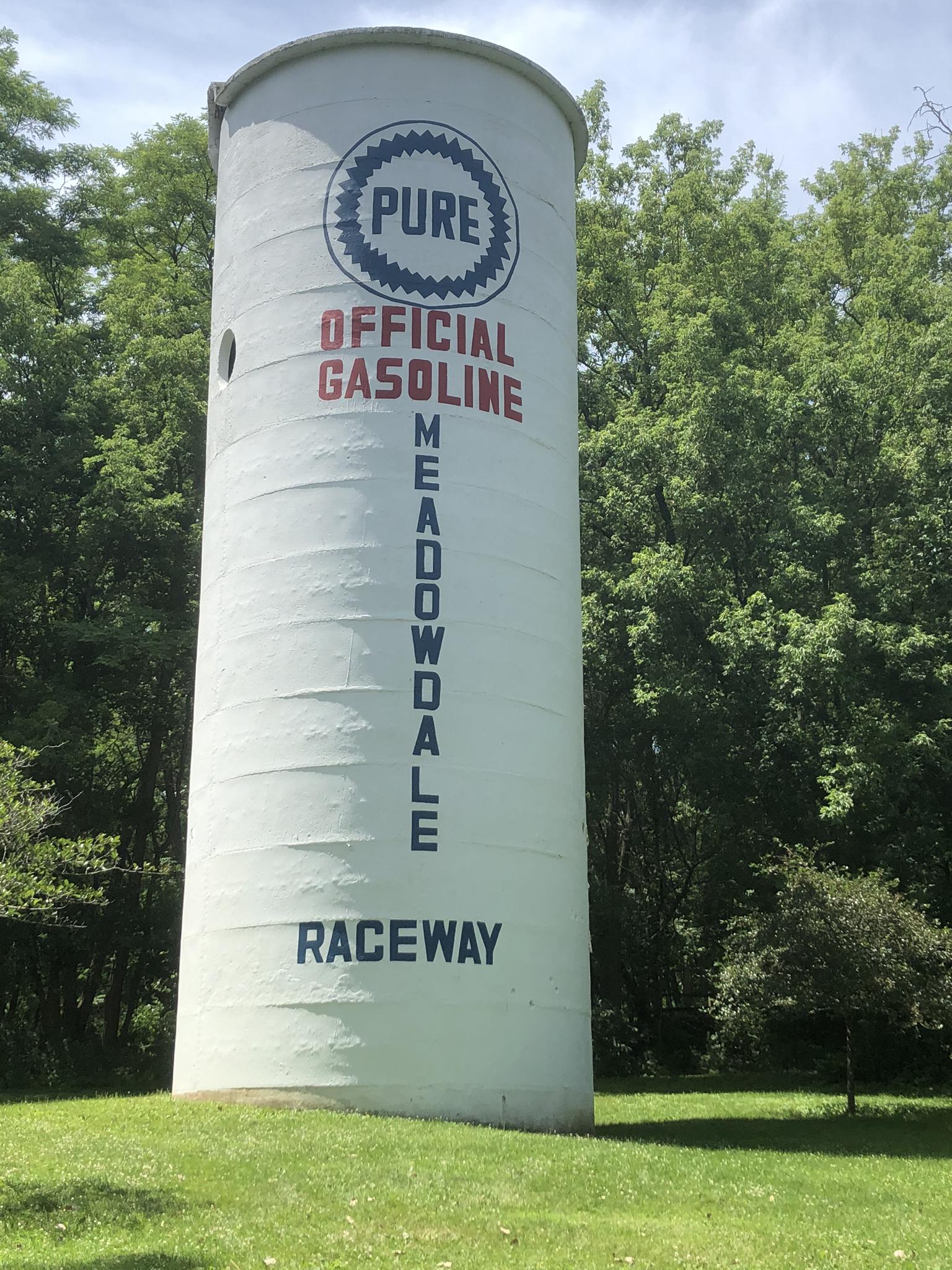 Tall white cylindrical structure with text, surrounded by green trees.