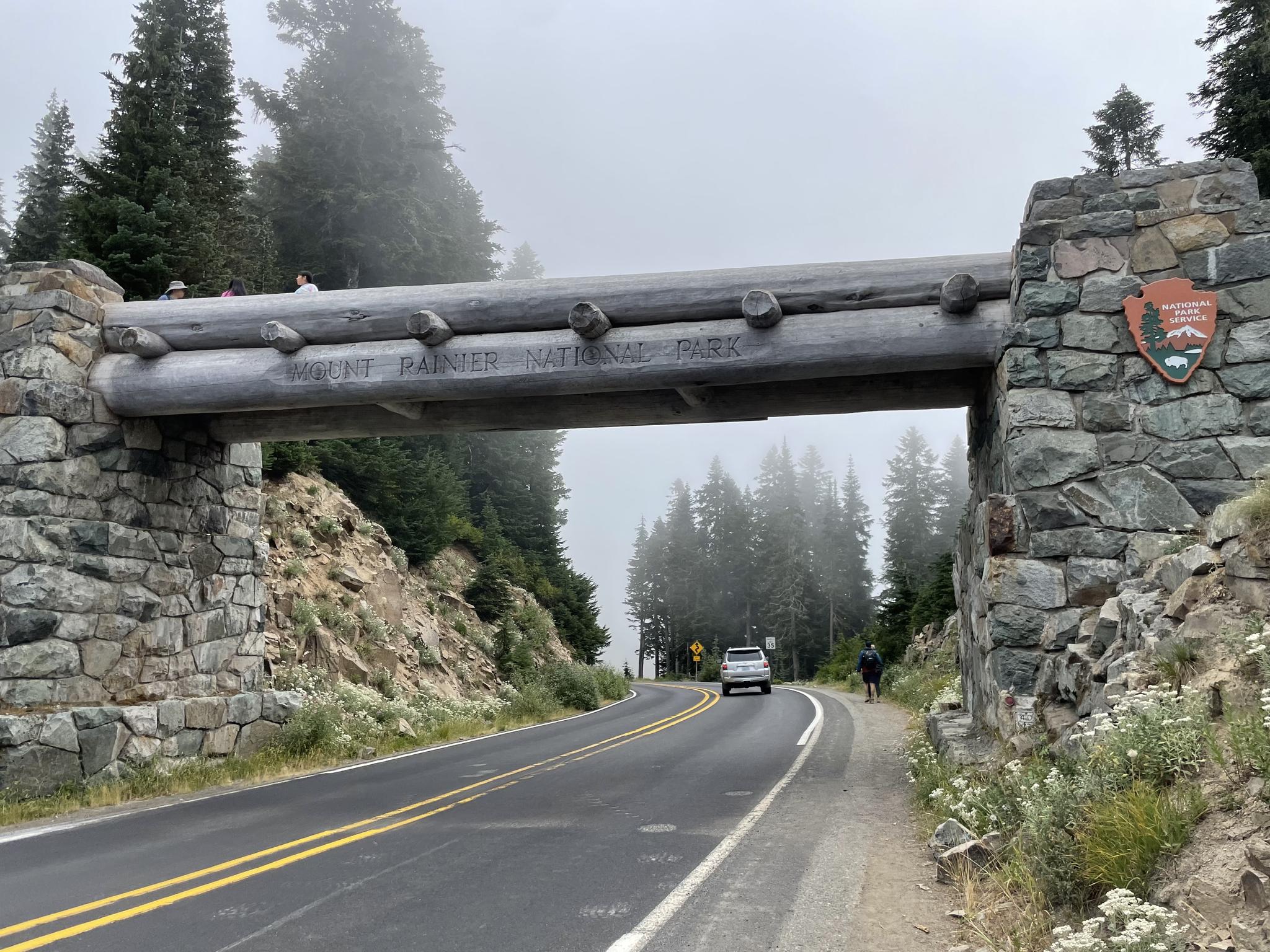 Road passing under a rustic log and stone archway on a foggy day.