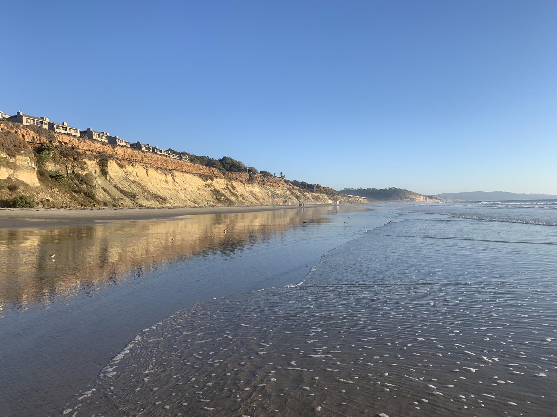 Sunny beach with gentle waves, tall cliffs, and distant houses under blue sky.