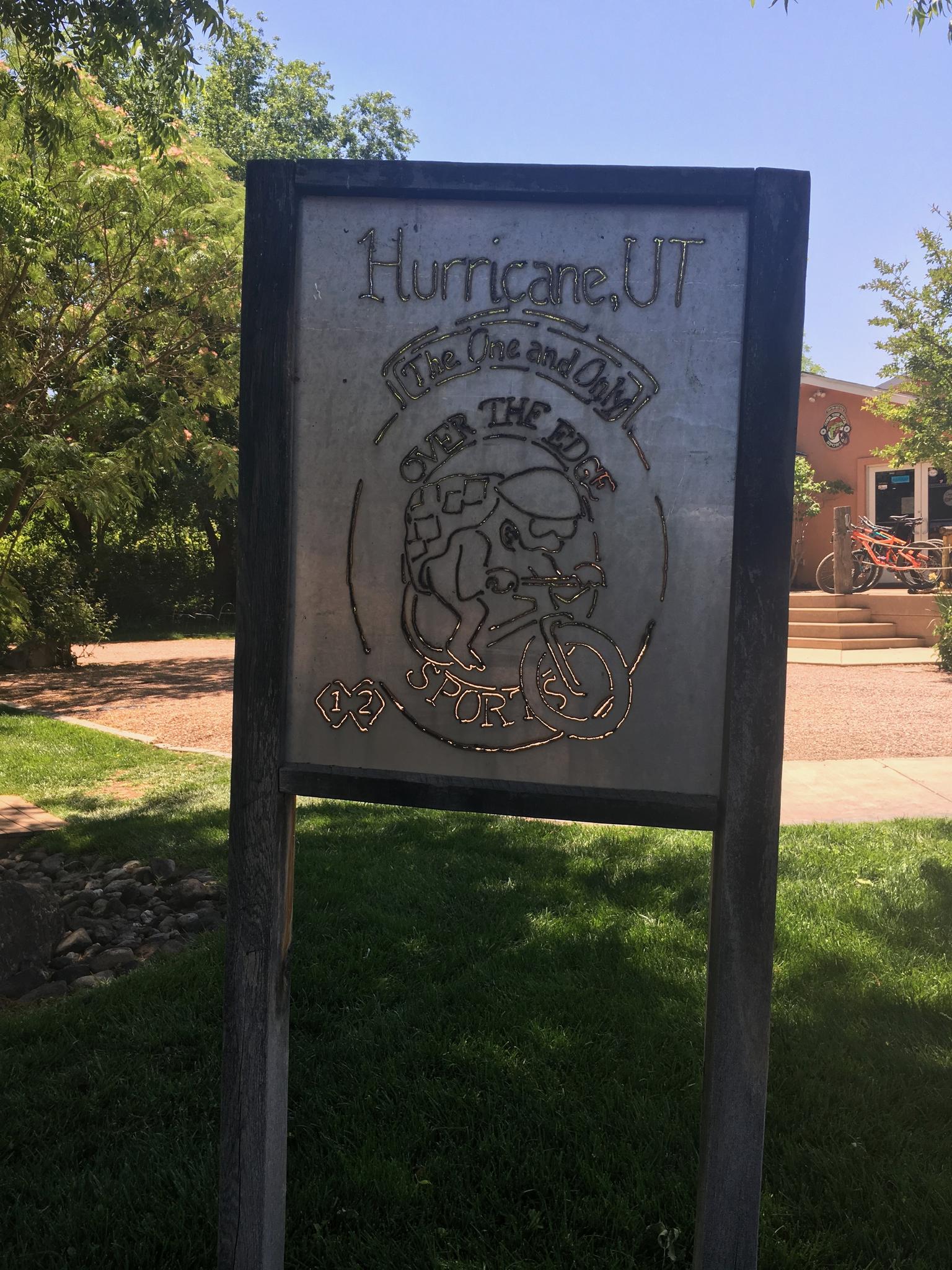 A weathered wooden sign with a bicycle graphic in a sunny, green park.
