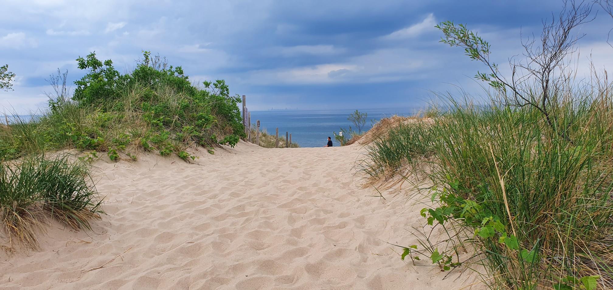 Sandy path through dunes with green grass leads to distant water and person.