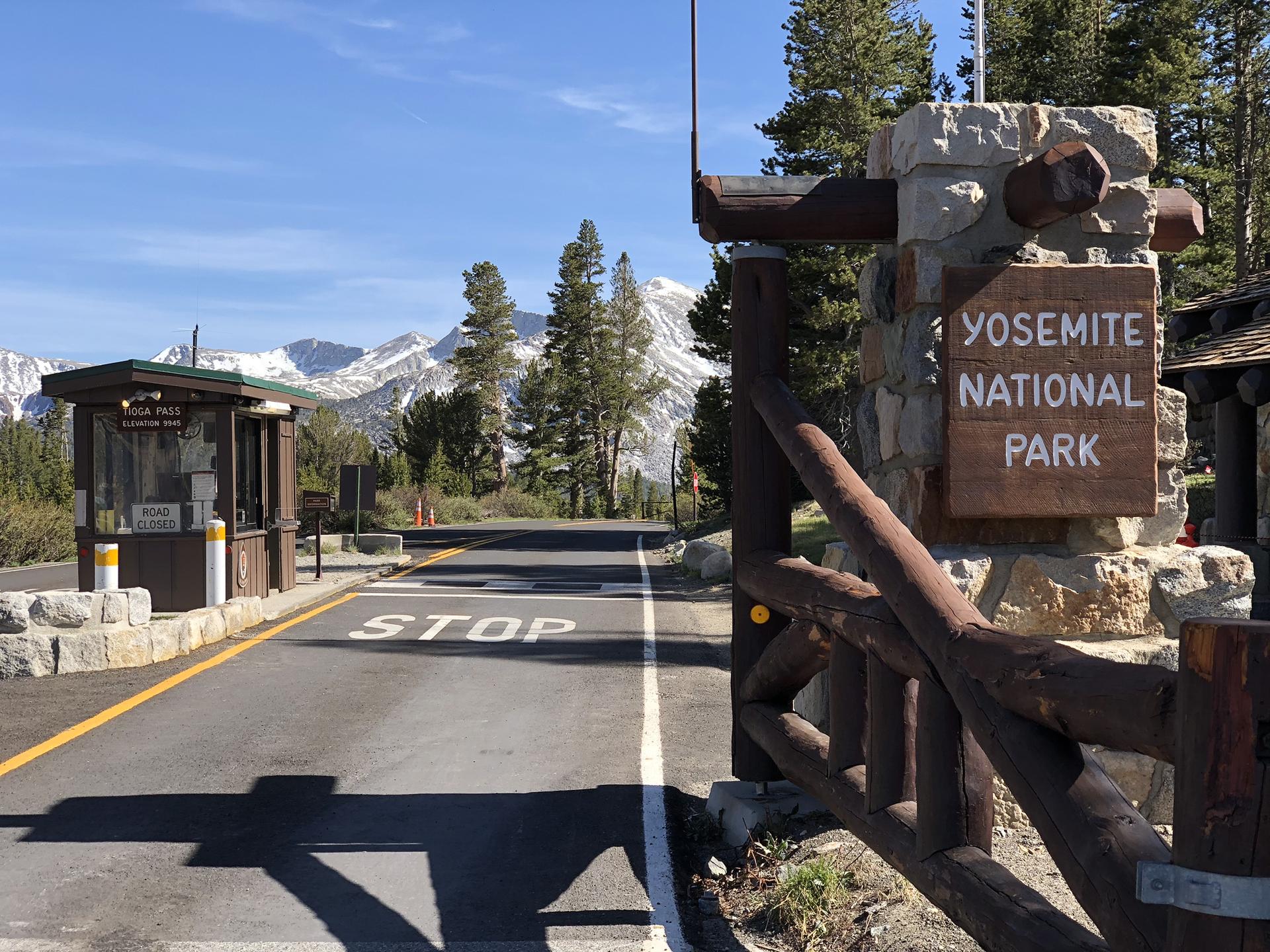 Road entrance with a wooden sign, mountains, and trees in bright daylight.