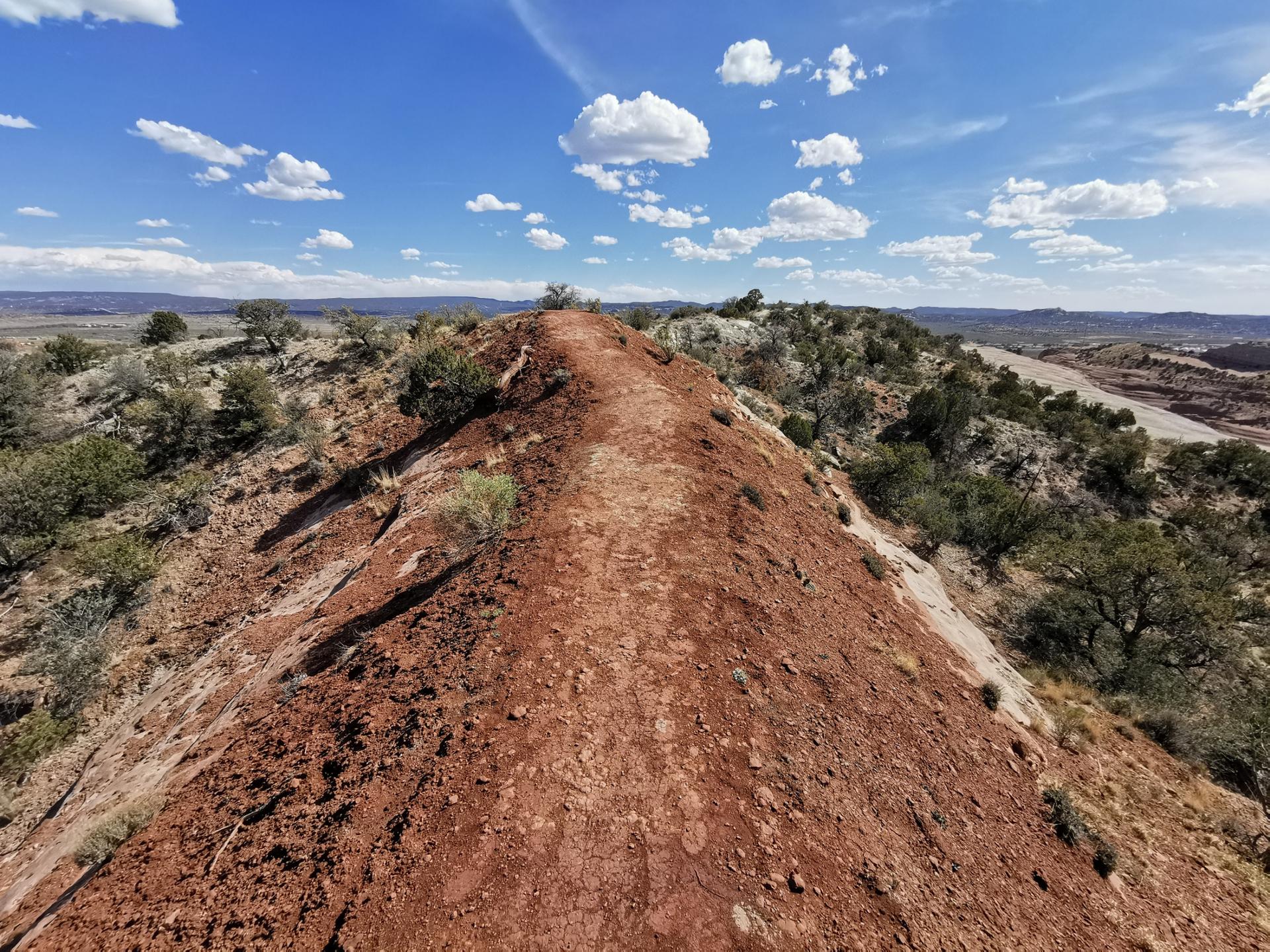A narrow dirt path winds along a red-earthed ridge under a bright, cloudy sky.