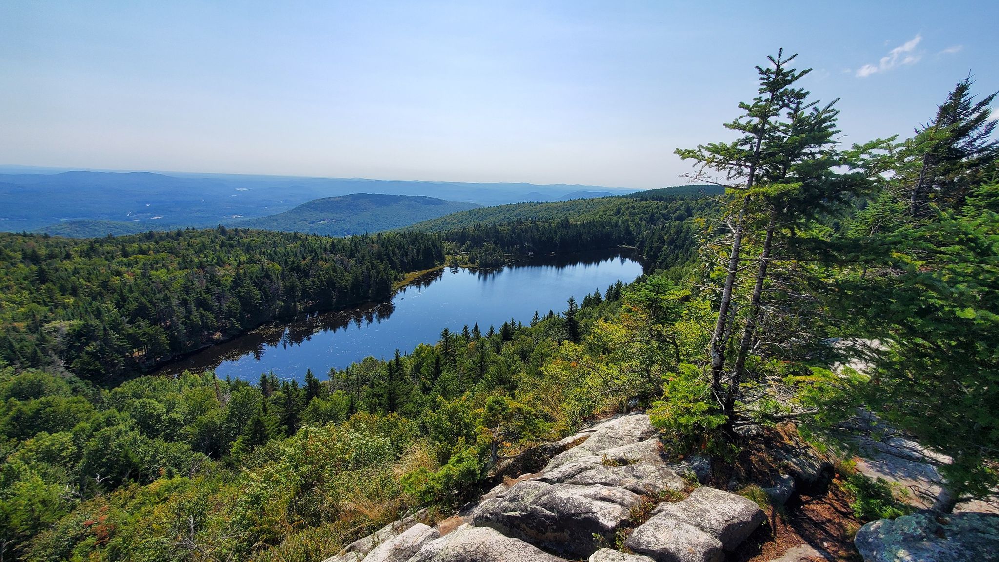 Sunny view of a calm lake surrounded by green forest from rocky outcrop. - Newbury Trail
