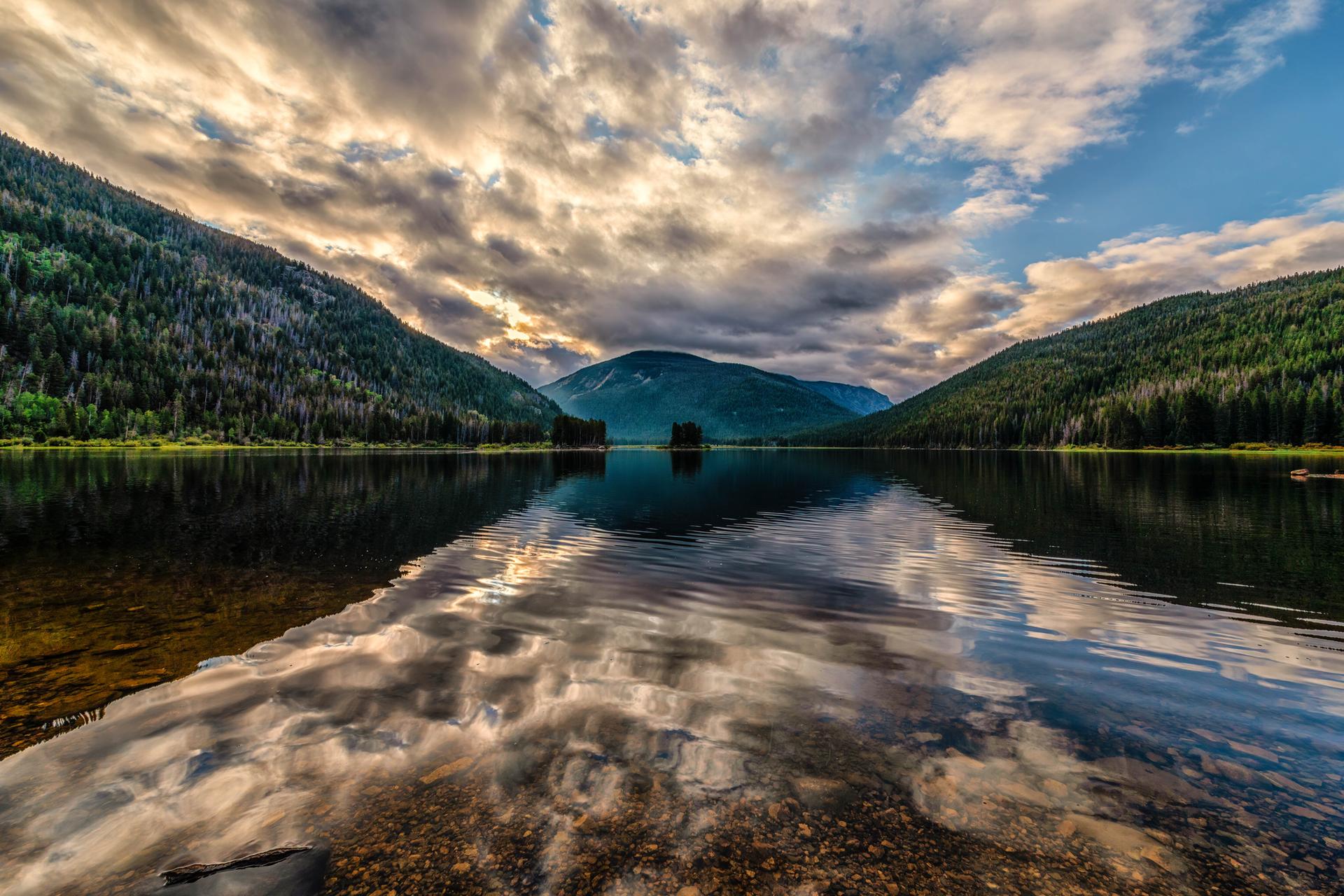 A serene lake reflects dramatic clouds and forested mountains at dusk.