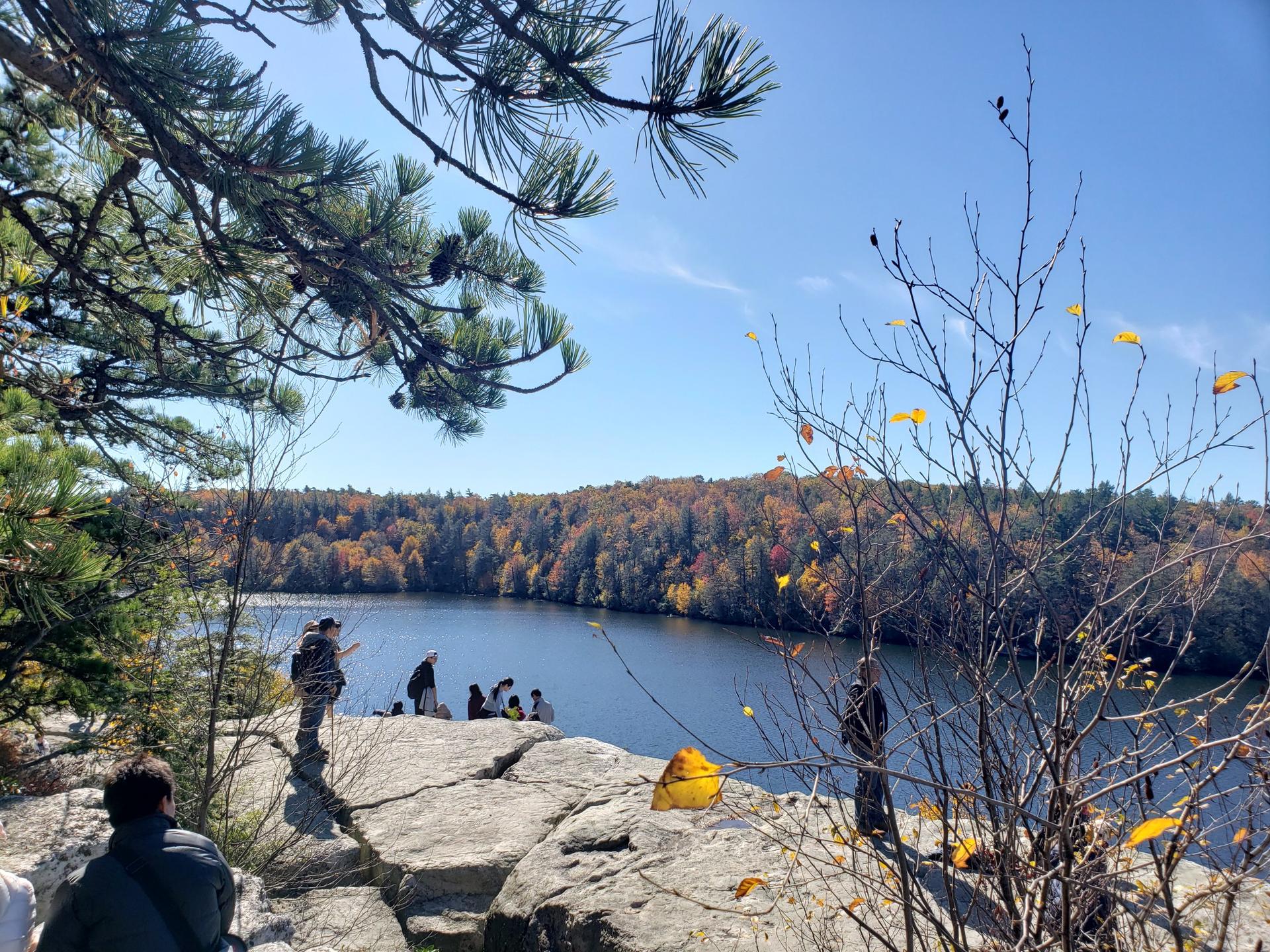 People on a rocky cliff overlooking a lake surrounded by autumn trees, sunny.