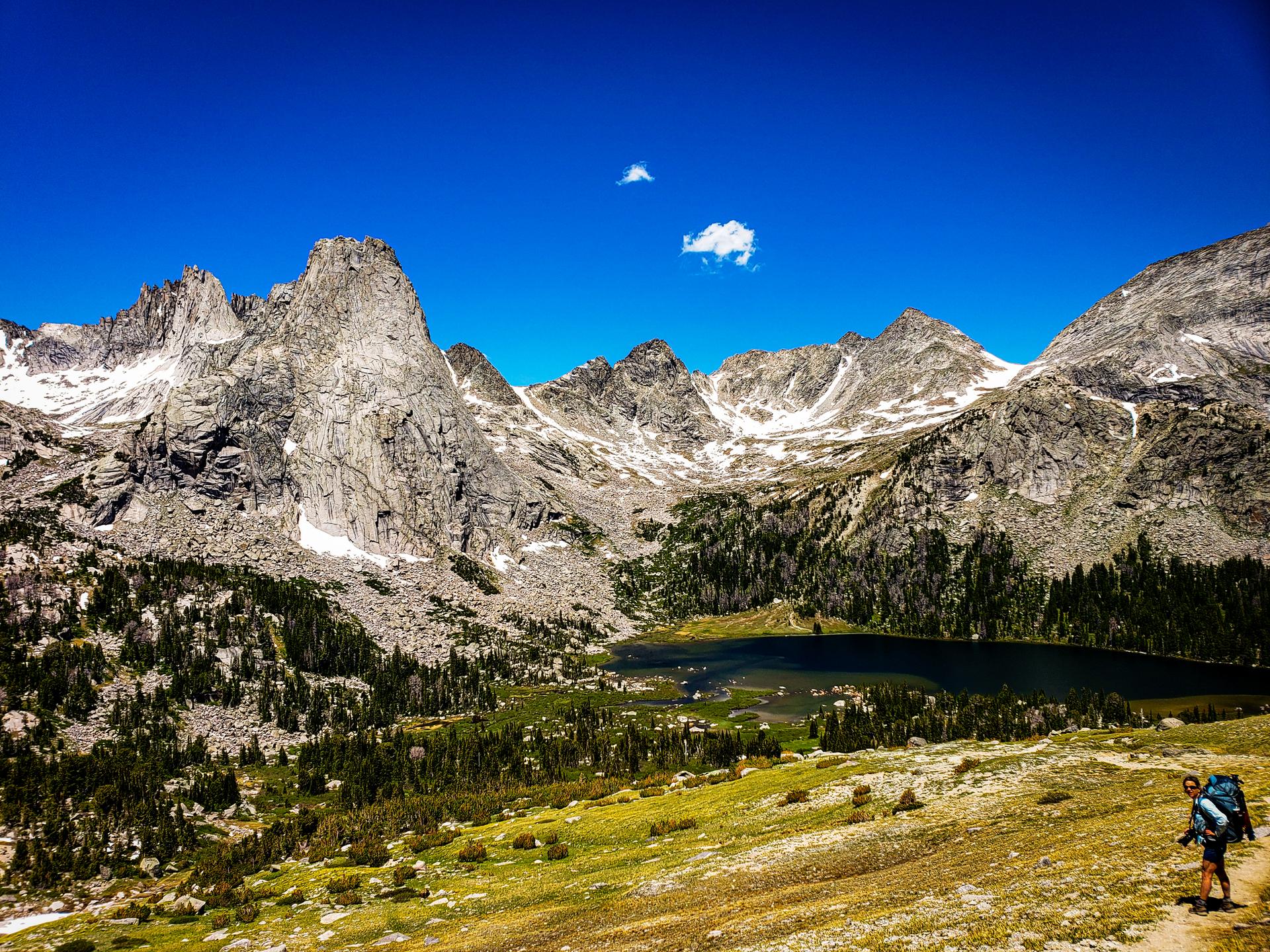A hiker walks a trail overlooking a vast mountain range and small lake.