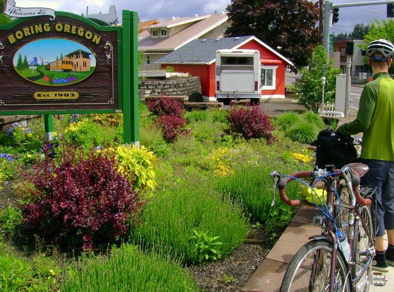 A person with a bicycle next to a sign and a colorful garden on a sunny day.