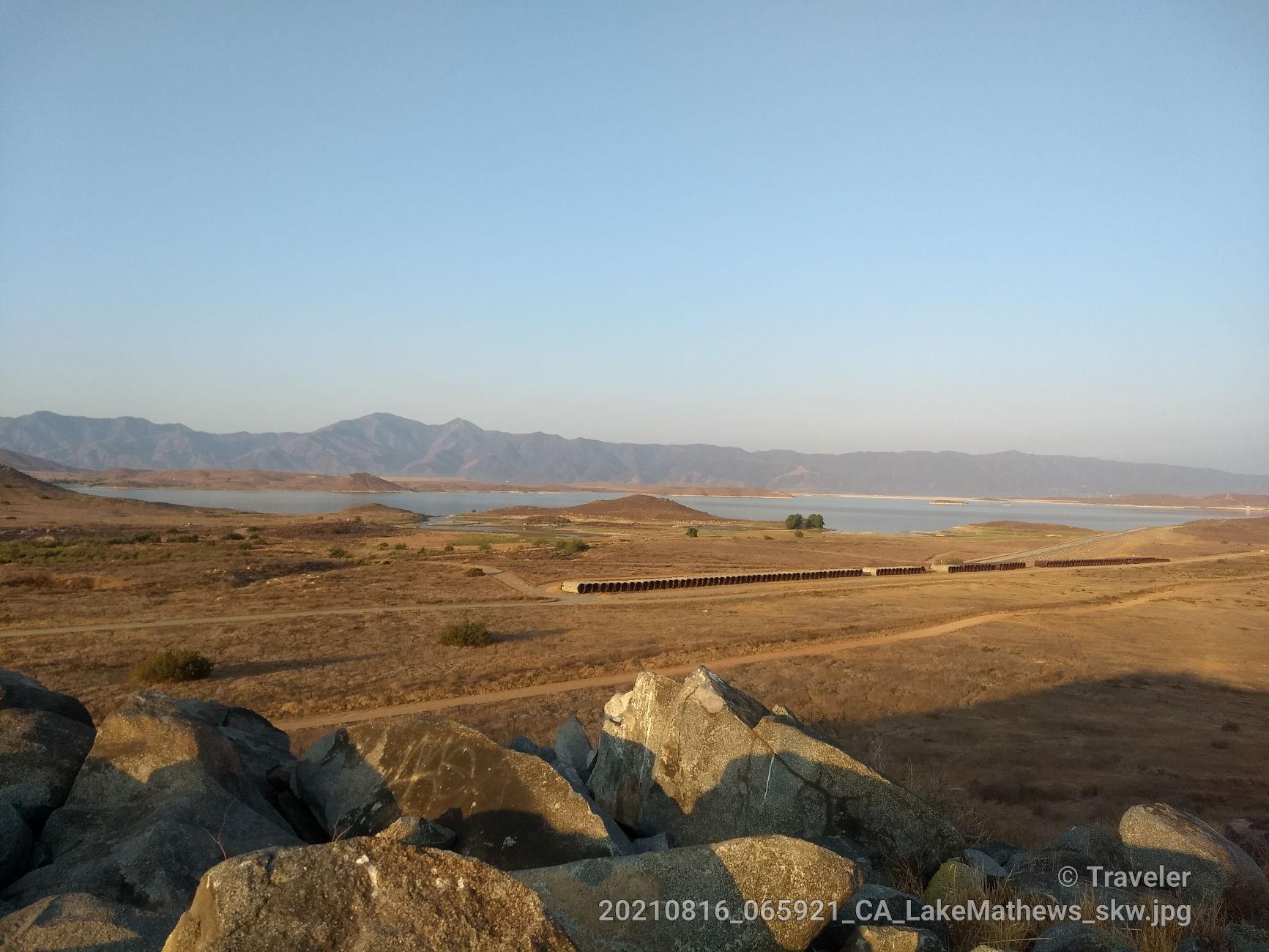 Sunny view of dry hills, a distant lake, mountains, and large foreground rocks.