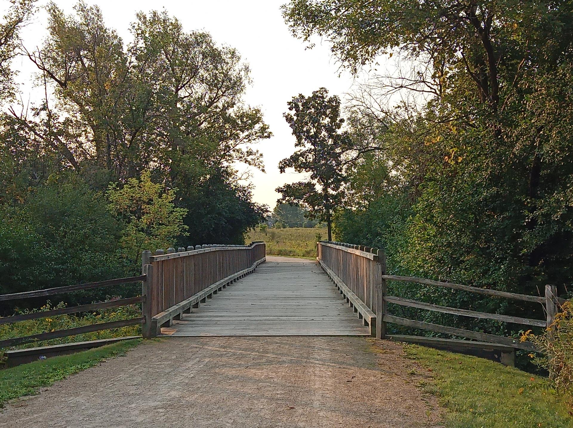 A wooden bridge with railings over a dirt path, leading into a field.