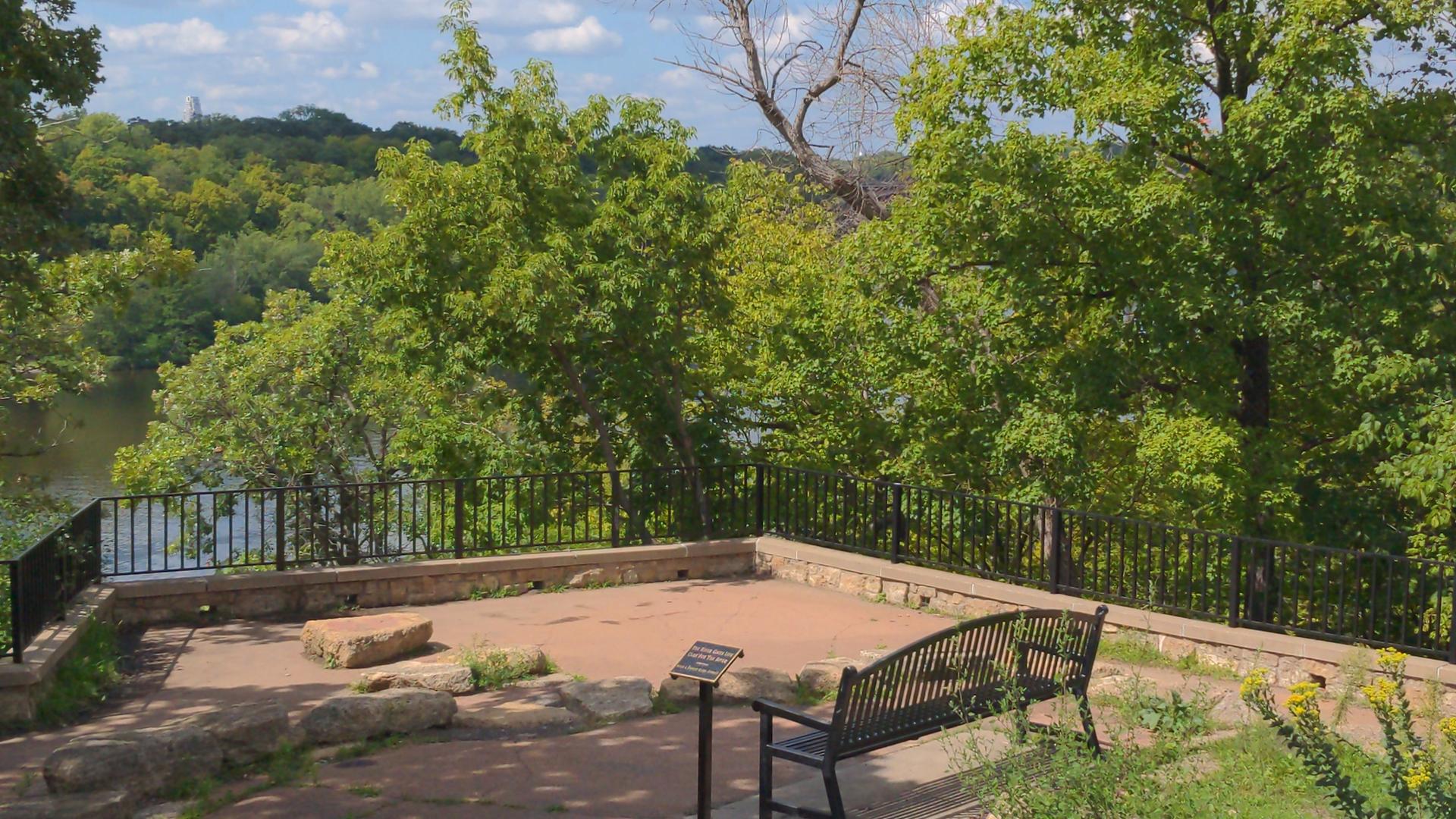 Sunny outdoor viewing platform with a bench overlooking a river and trees.