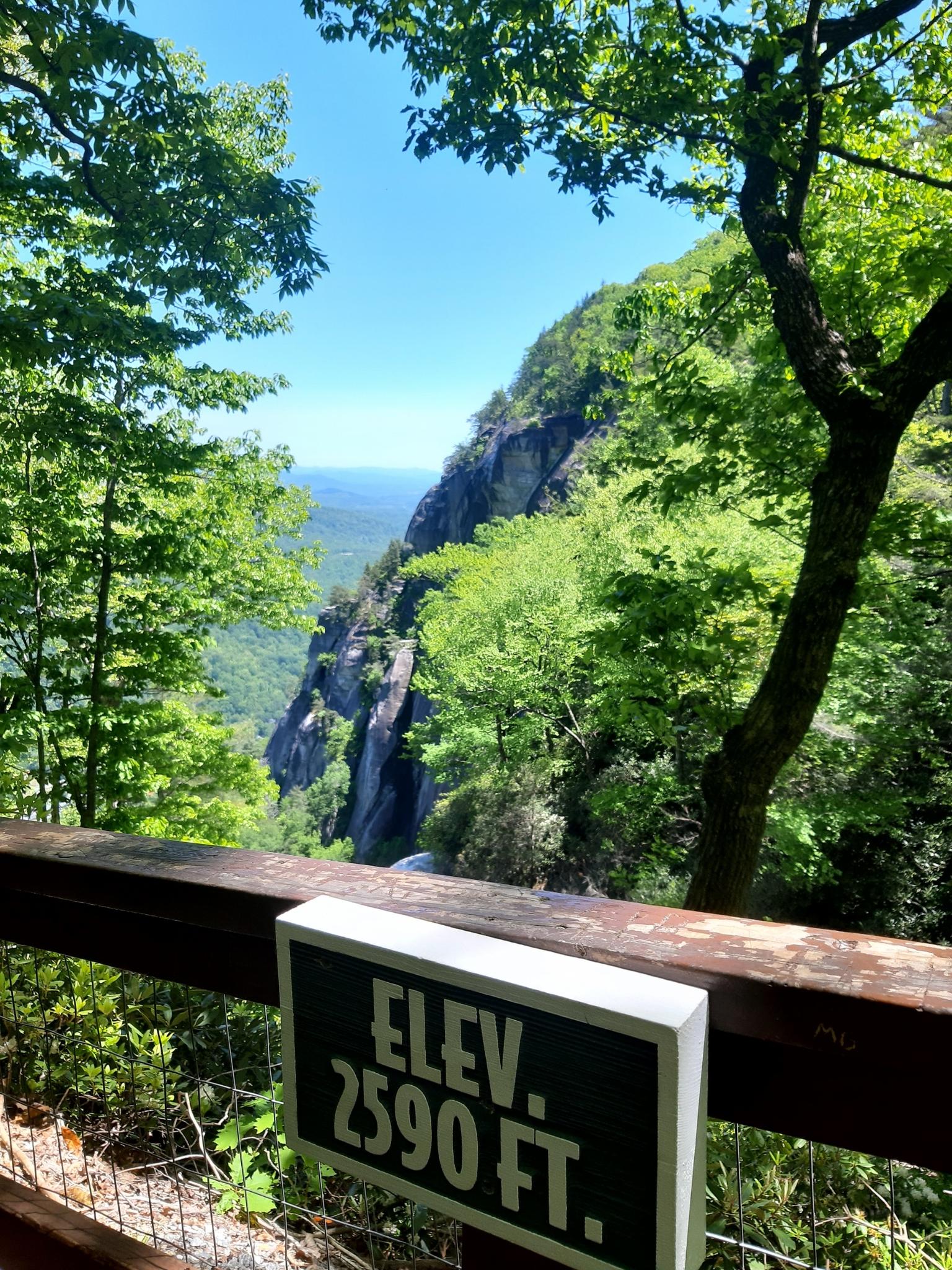 Wooden railing with an "ELEV 2590 FT" sign, overlooking a rocky cliff and valley. Bright daylight.