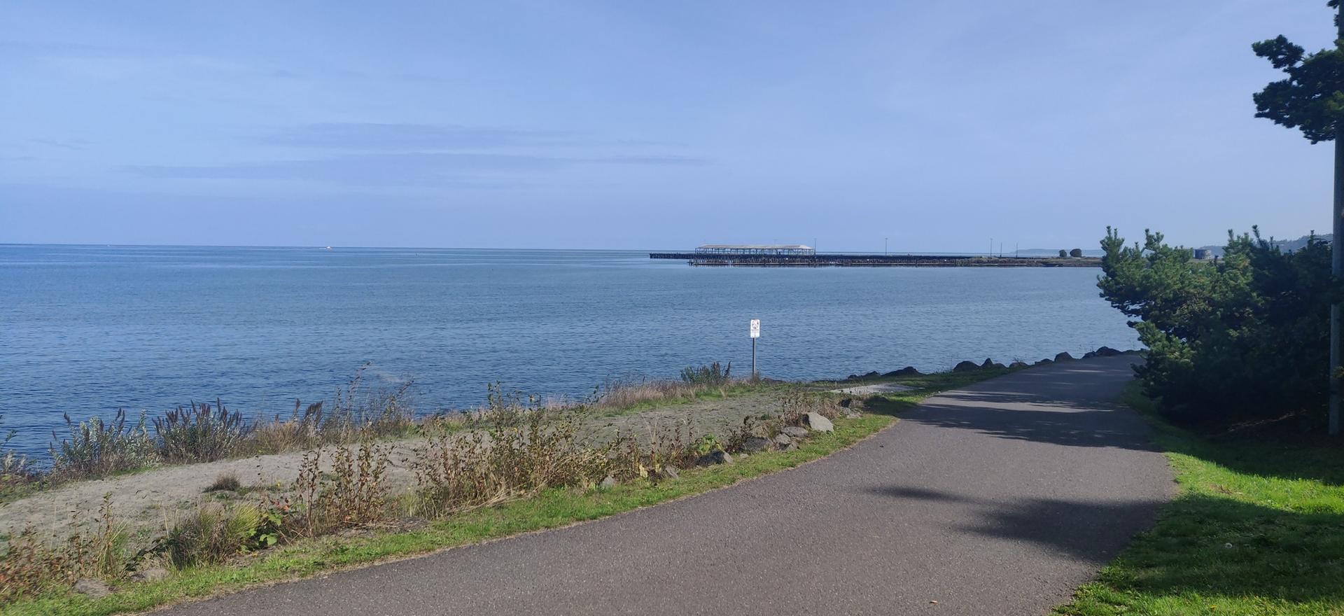 A paved path runs alongside a calm blue ocean under a clear sky.