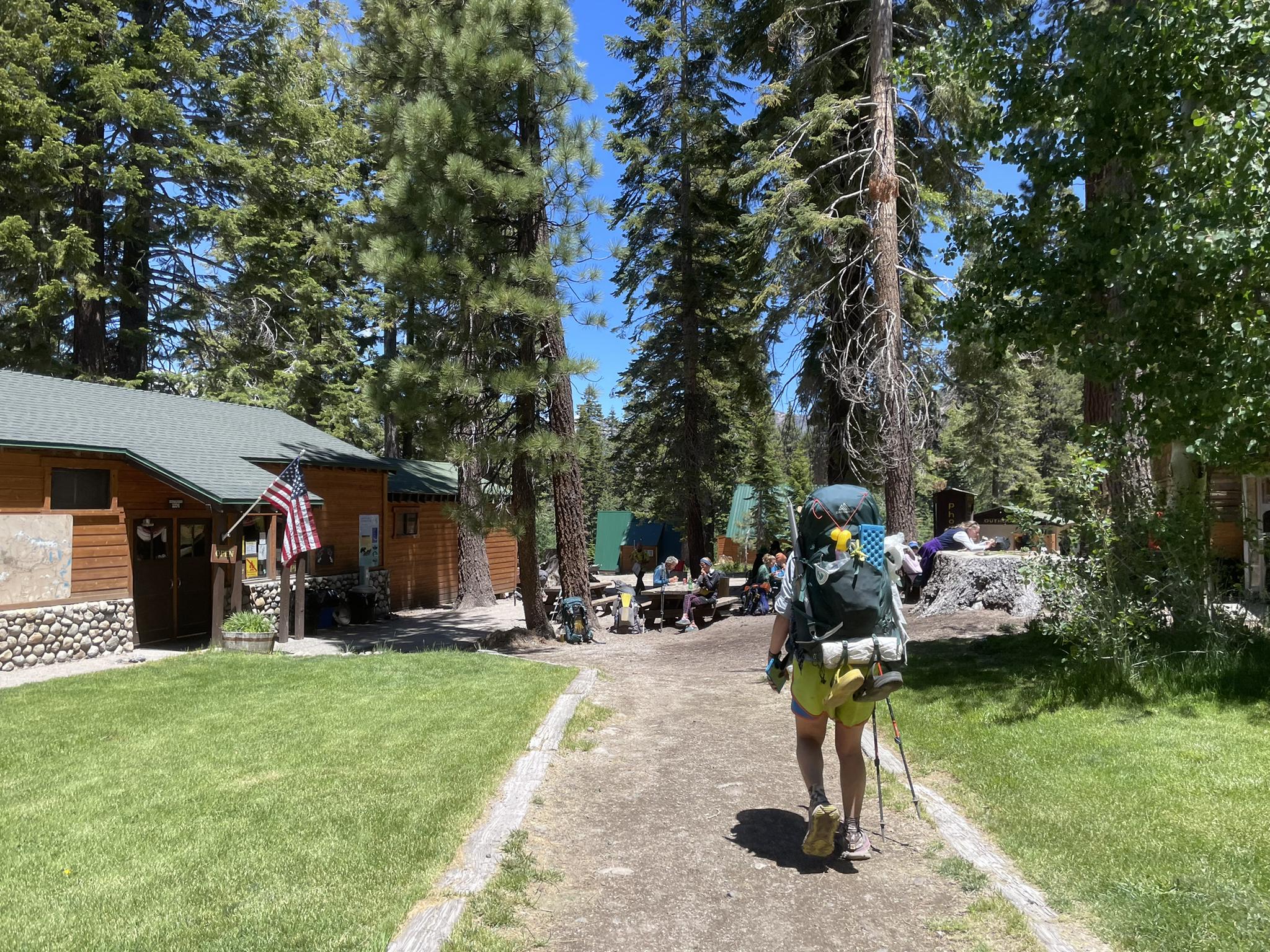 A person with a large backpack walks on a path past cabins and tall pine trees.