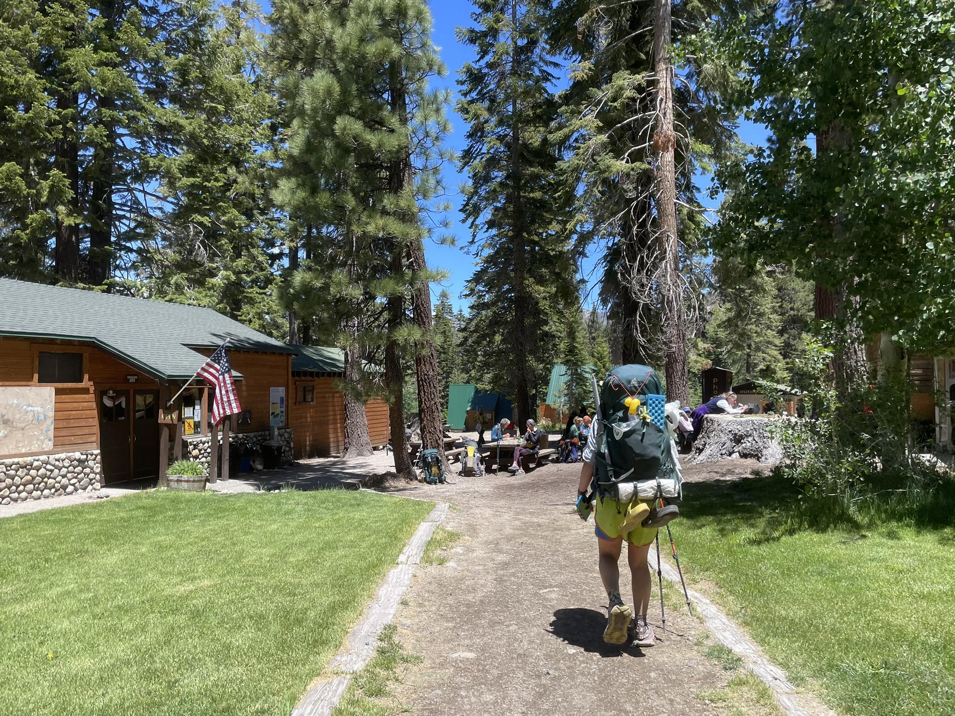 A person with a large backpack walks on a path past cabins and tall pine trees.