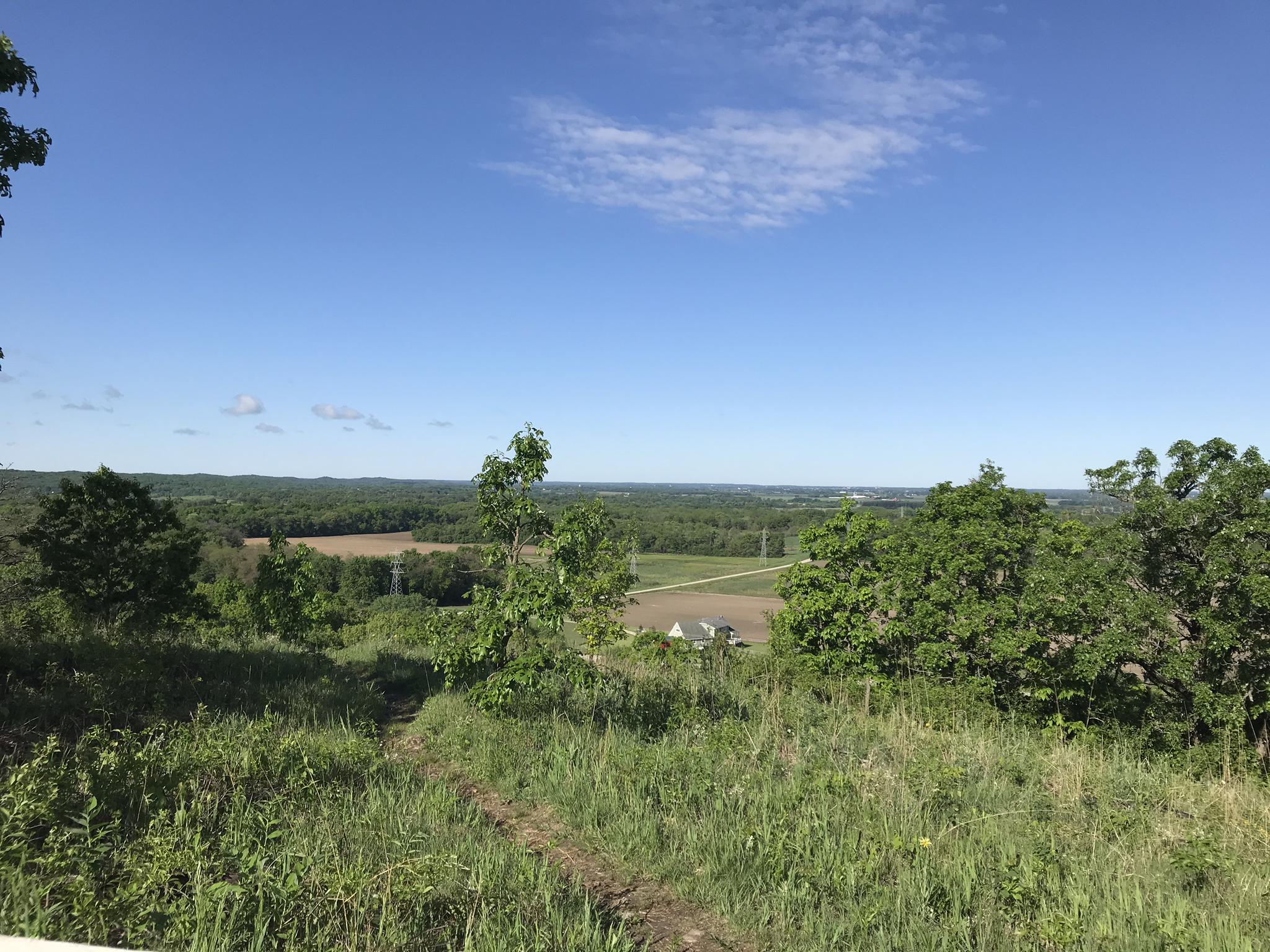 Sunny overlook of a green valley with distant fields and a clear sky.