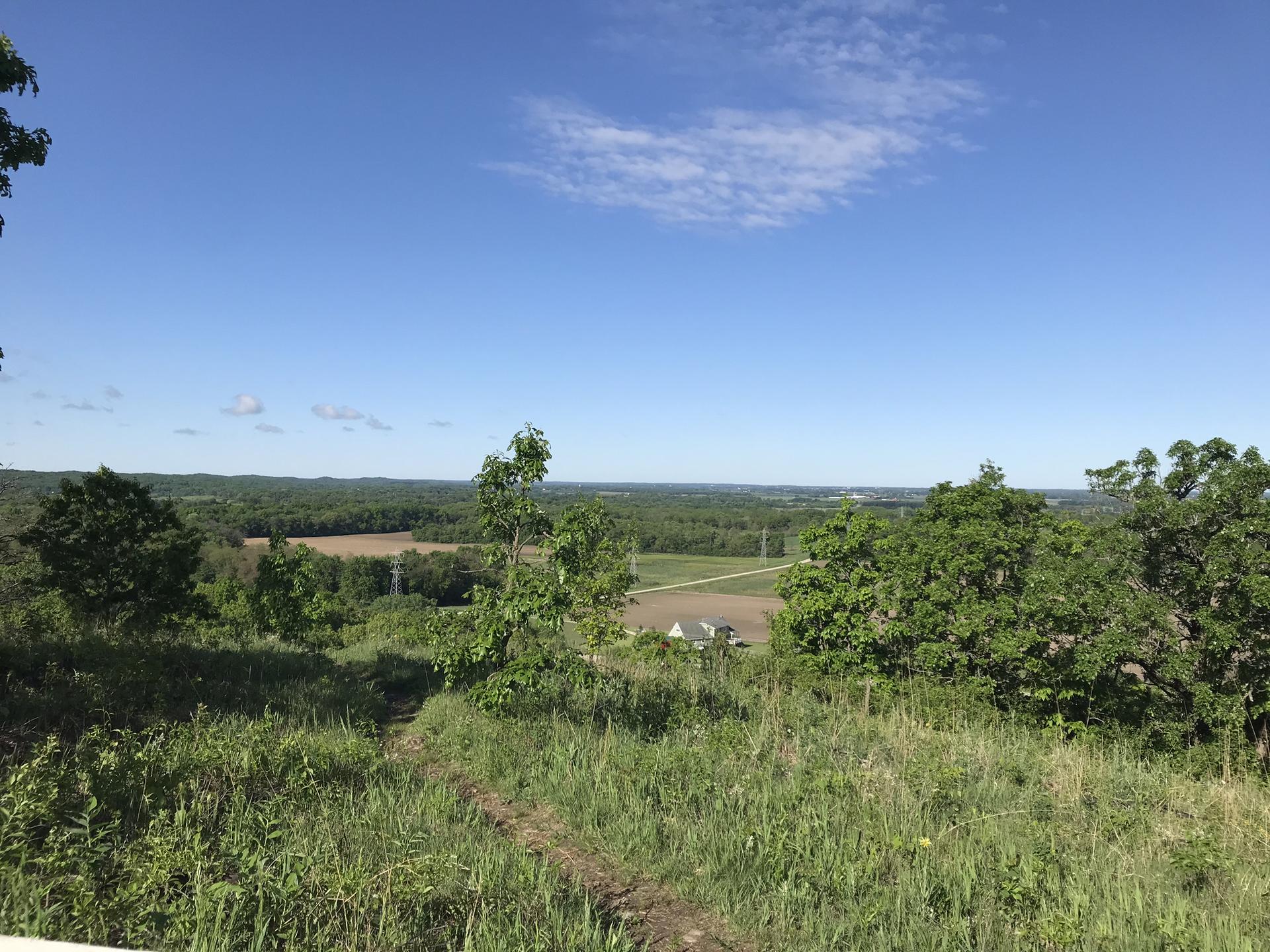 Sunny overlook of a green valley with distant fields and a clear sky.