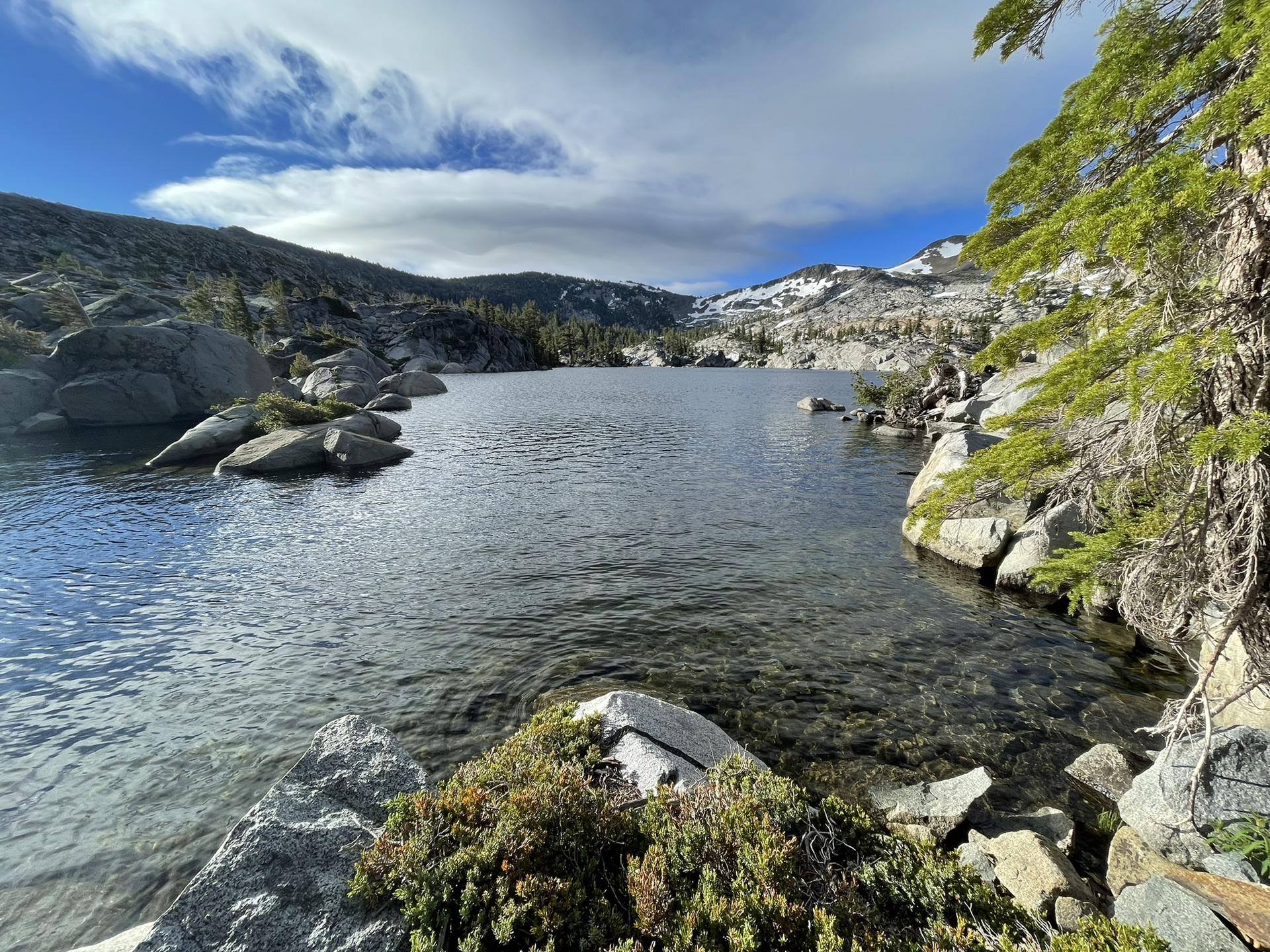 Clear lake with rocky shores, pine trees, and snow-capped mountains.