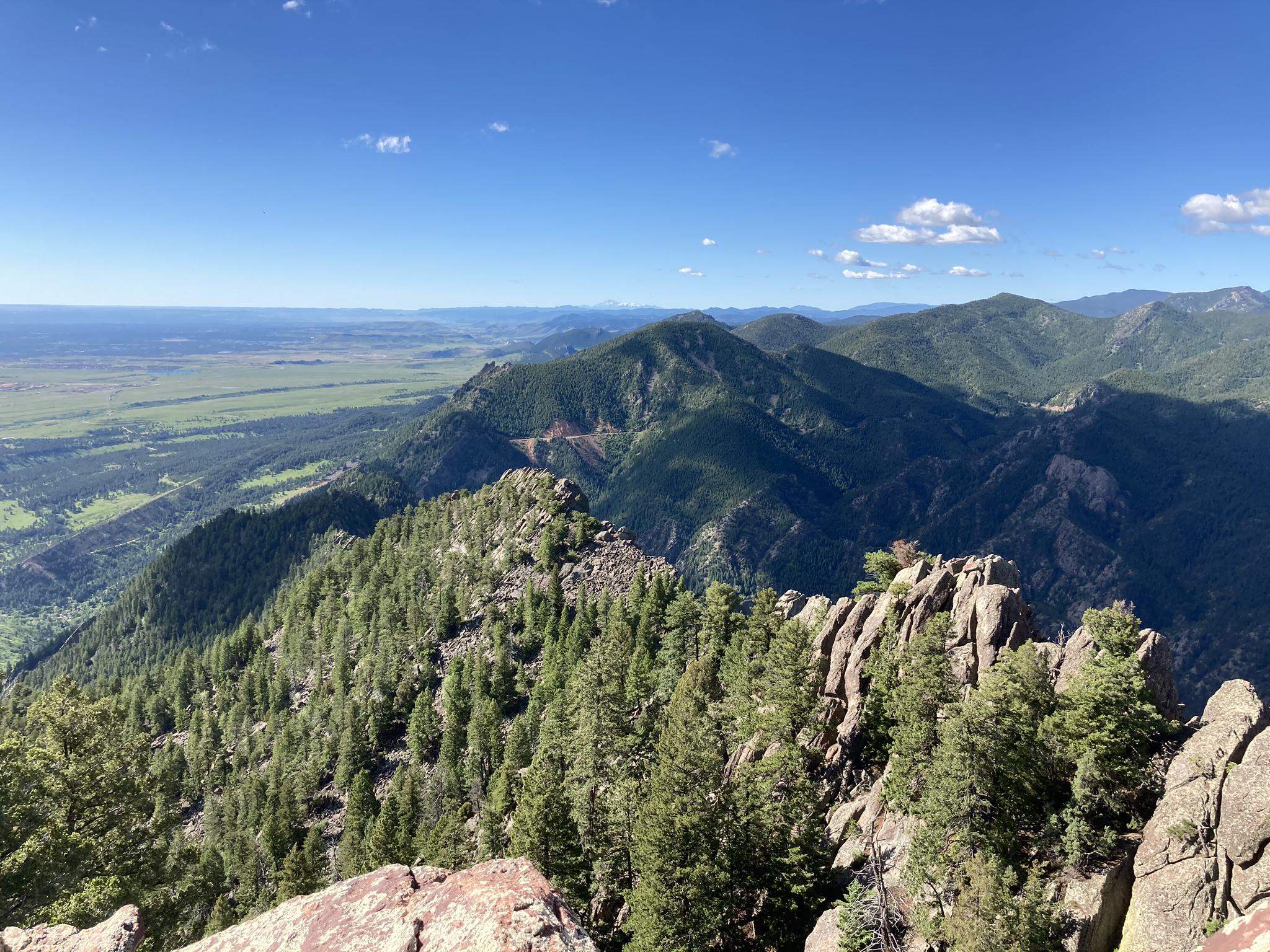 A forested mountain ridge with rocky outcrops and a vast plain under blue sky.