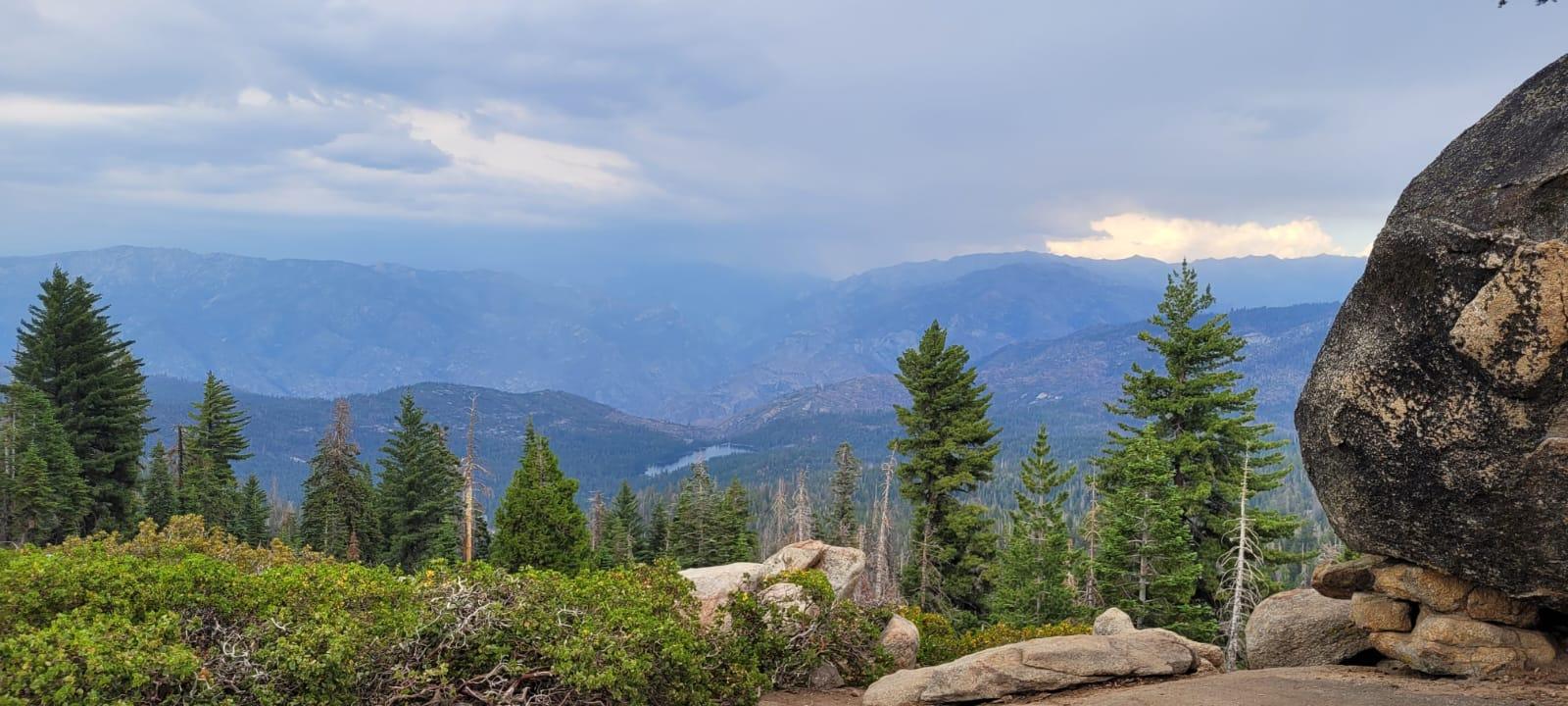 Panoramic mountain vista with a valley and pine trees under a cloudy sky.