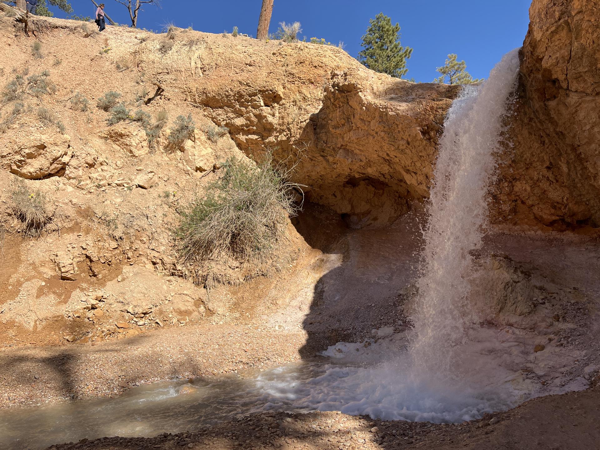 Waterfall cascading over a red rock cliff into a stream, person visible above.
