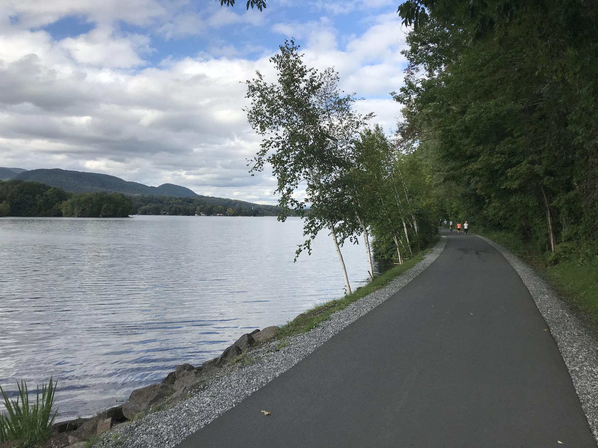 Paved path alongside a lake with distant mountains under cloudy sky.