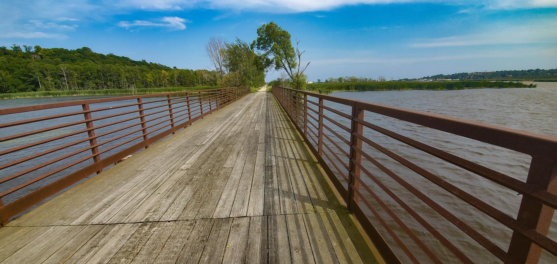 Sunny wooden bridge over water with railings and distant green hills.