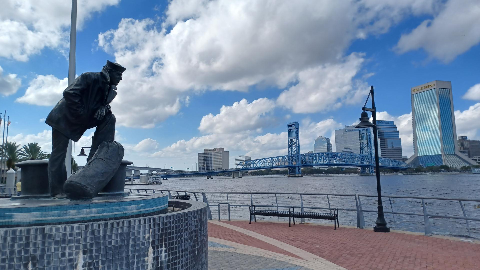 Statue of a man overlooking a river, blue bridge, and city skyline.