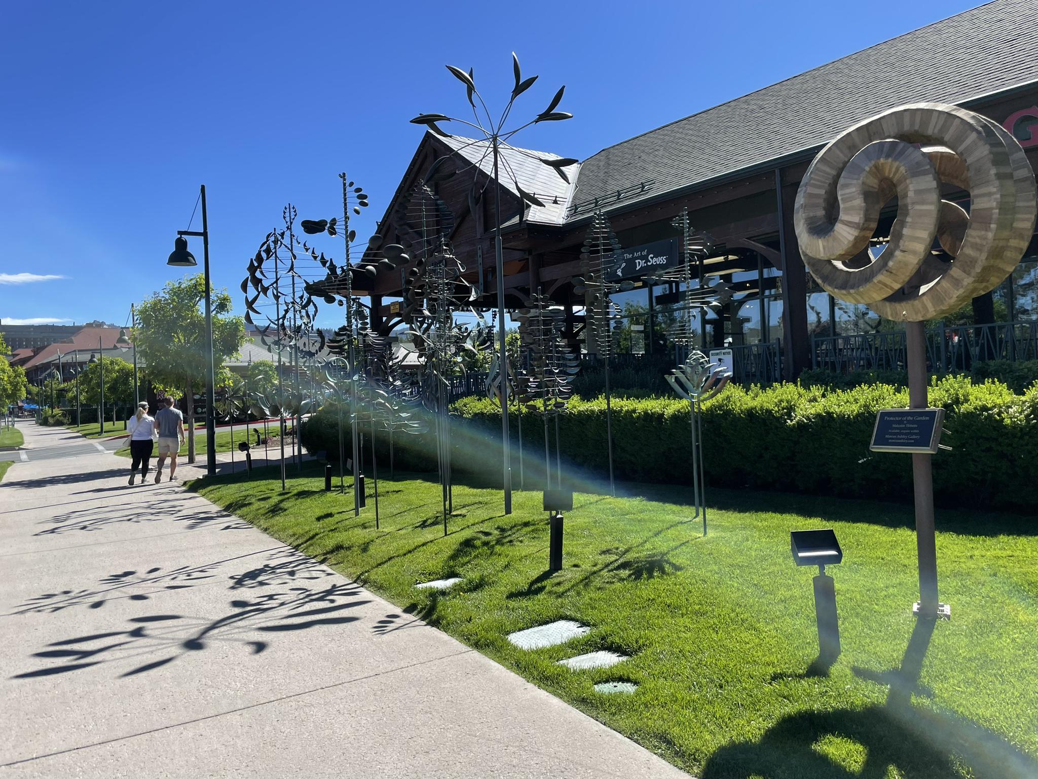 People walk past kinetic sculptures and a building on a sunny day.