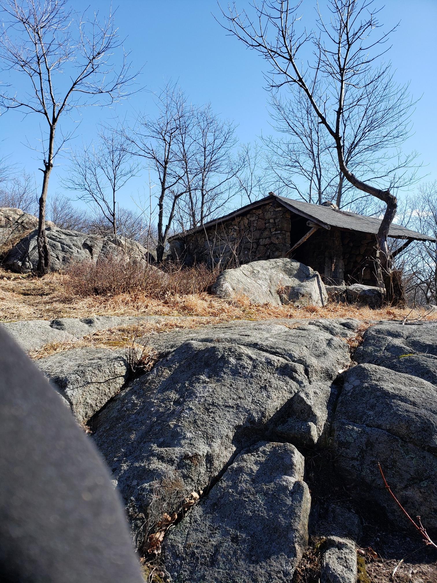 Rustic stone cabin nestled among large rocks and bare trees on a sunny day.