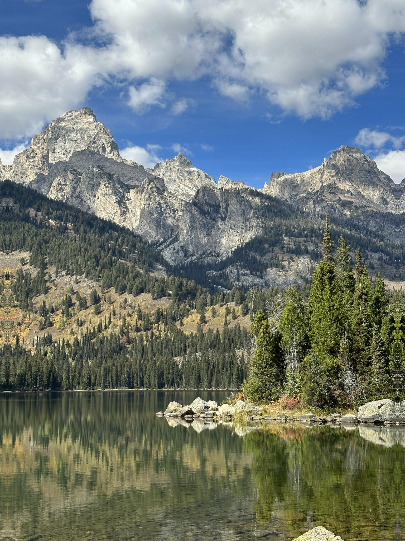 Clear lake reflecting rugged mountains and pine trees under a blue sky.
