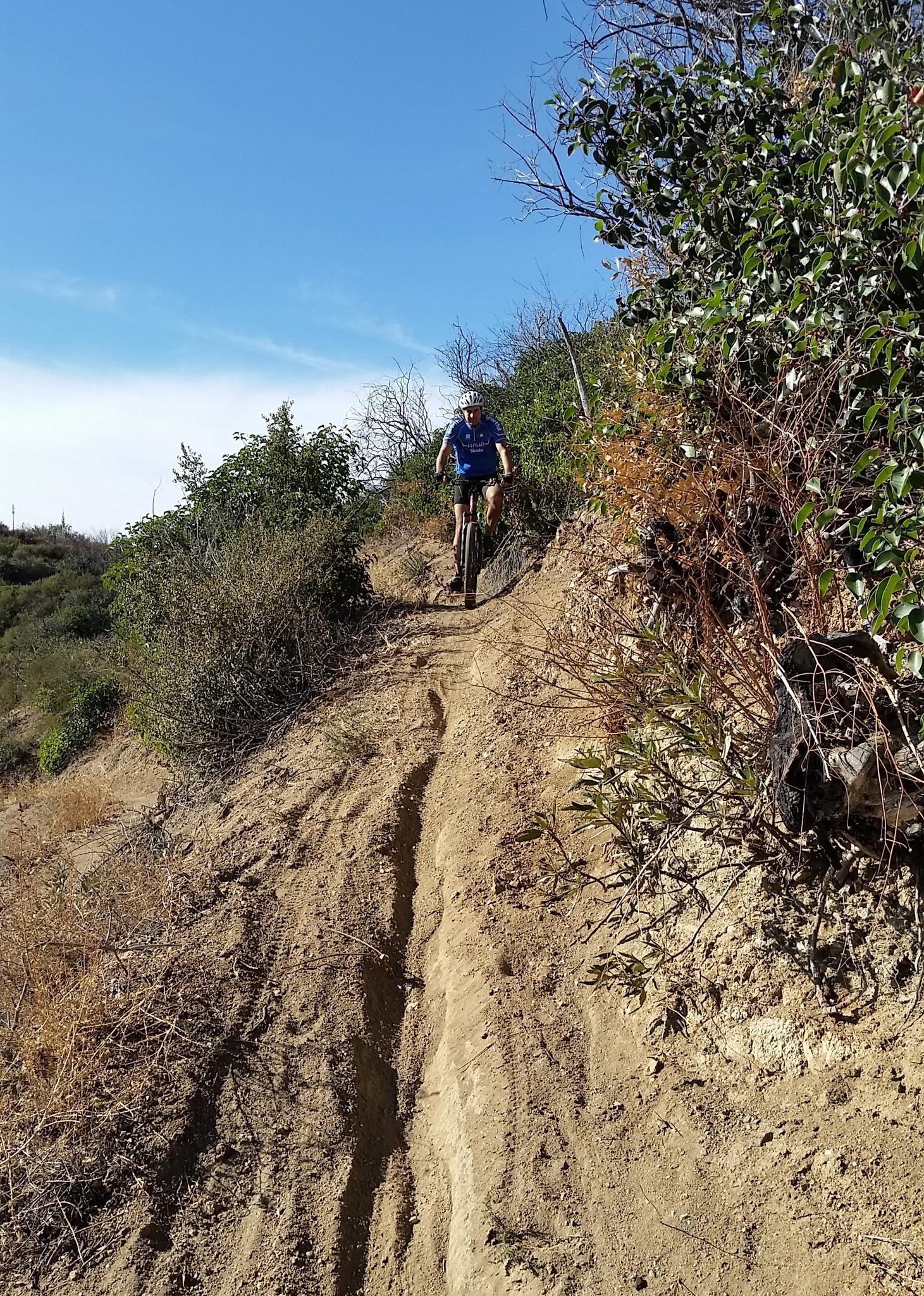 A mountain biker descends a dry, steep dirt path under a clear sky.