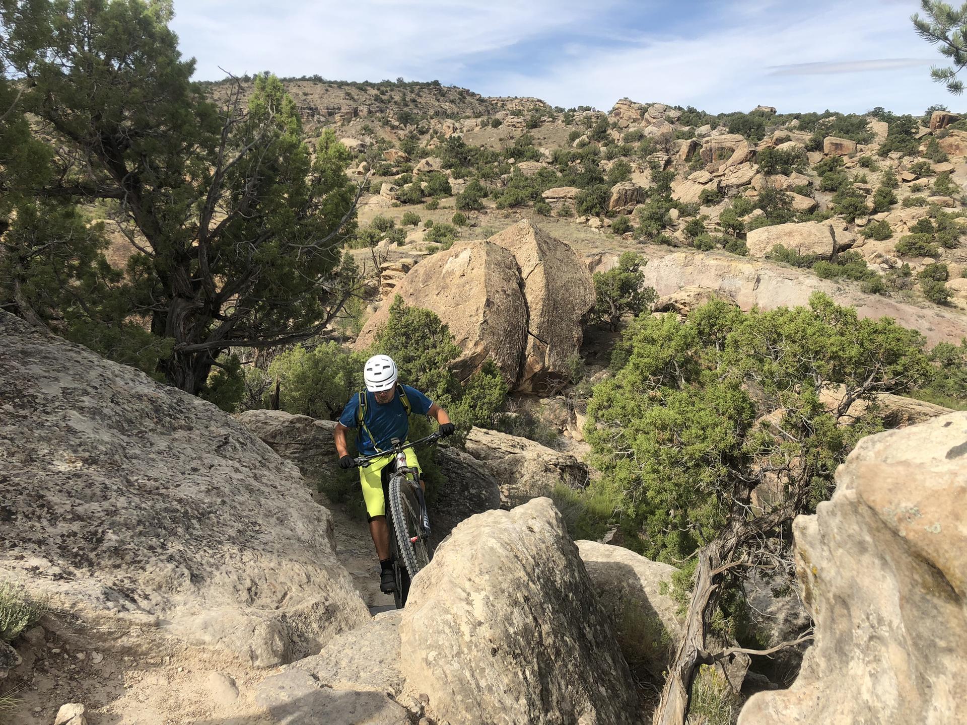 A mountain biker navigates a rocky, technical trail through arid, boulder-strewn terrain.