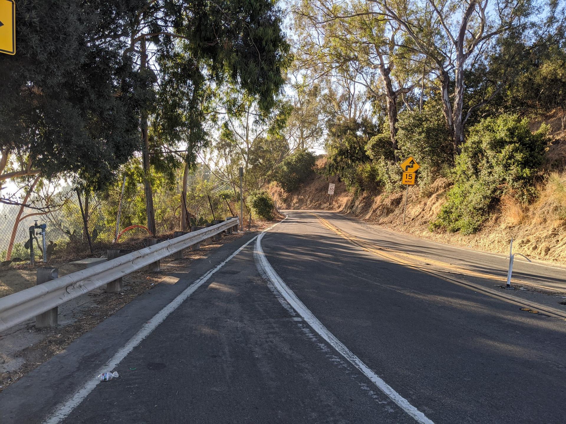 A winding road with a guardrail through sunlit trees.