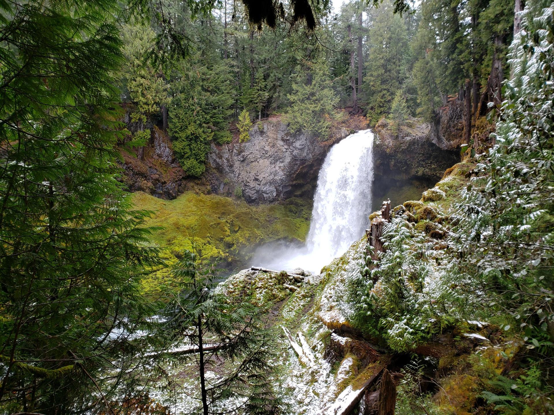Waterfall plunges into a river, surrounded by snowy evergreen forest on an overcast day.