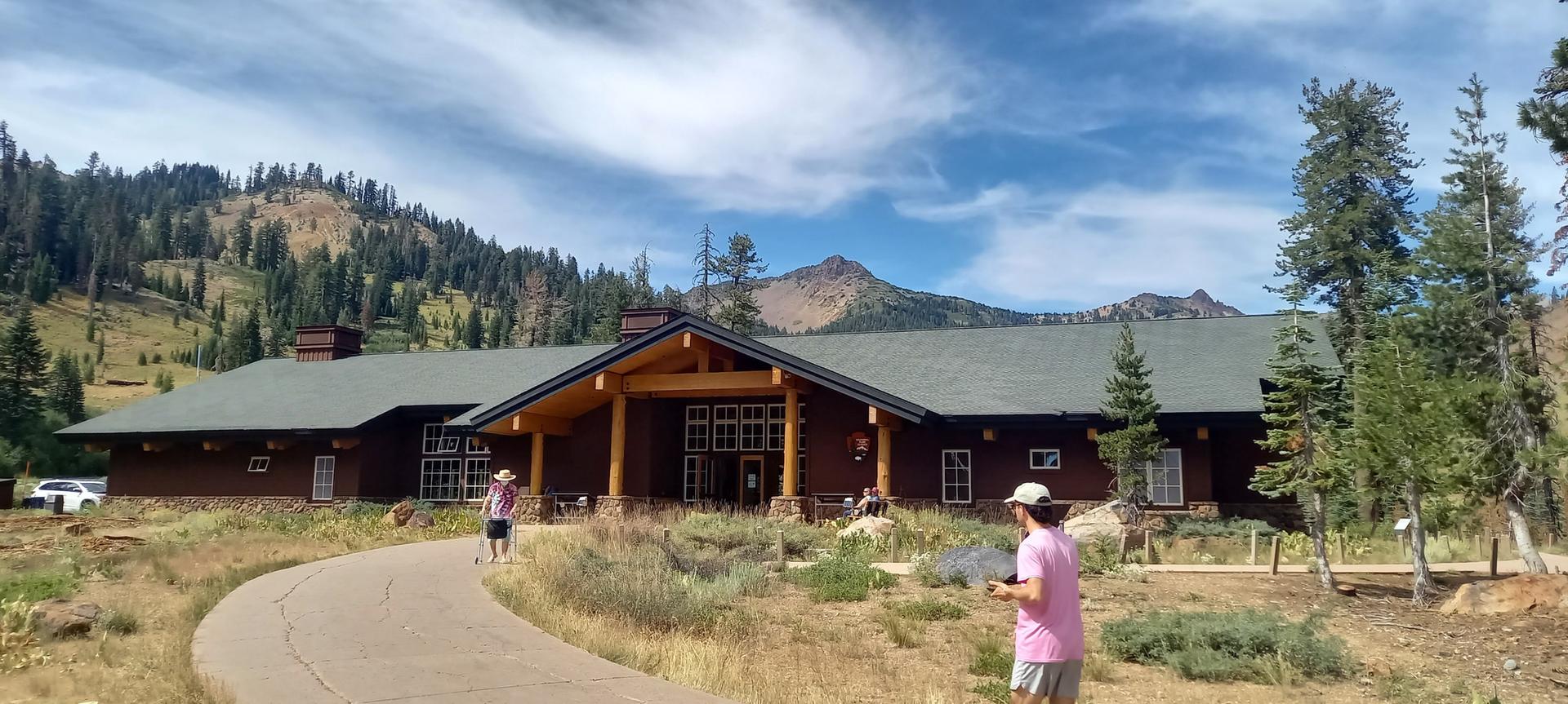 People walk on a paved path towards a large wooden building with mountains.