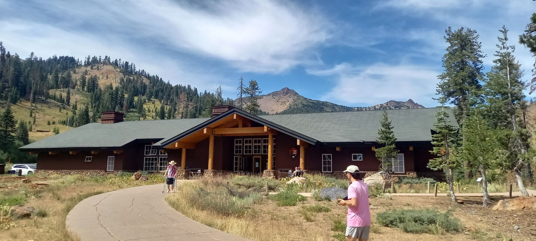 People walk on a paved path towards a large wooden building with mountains. - Mill Creek Falls Trail