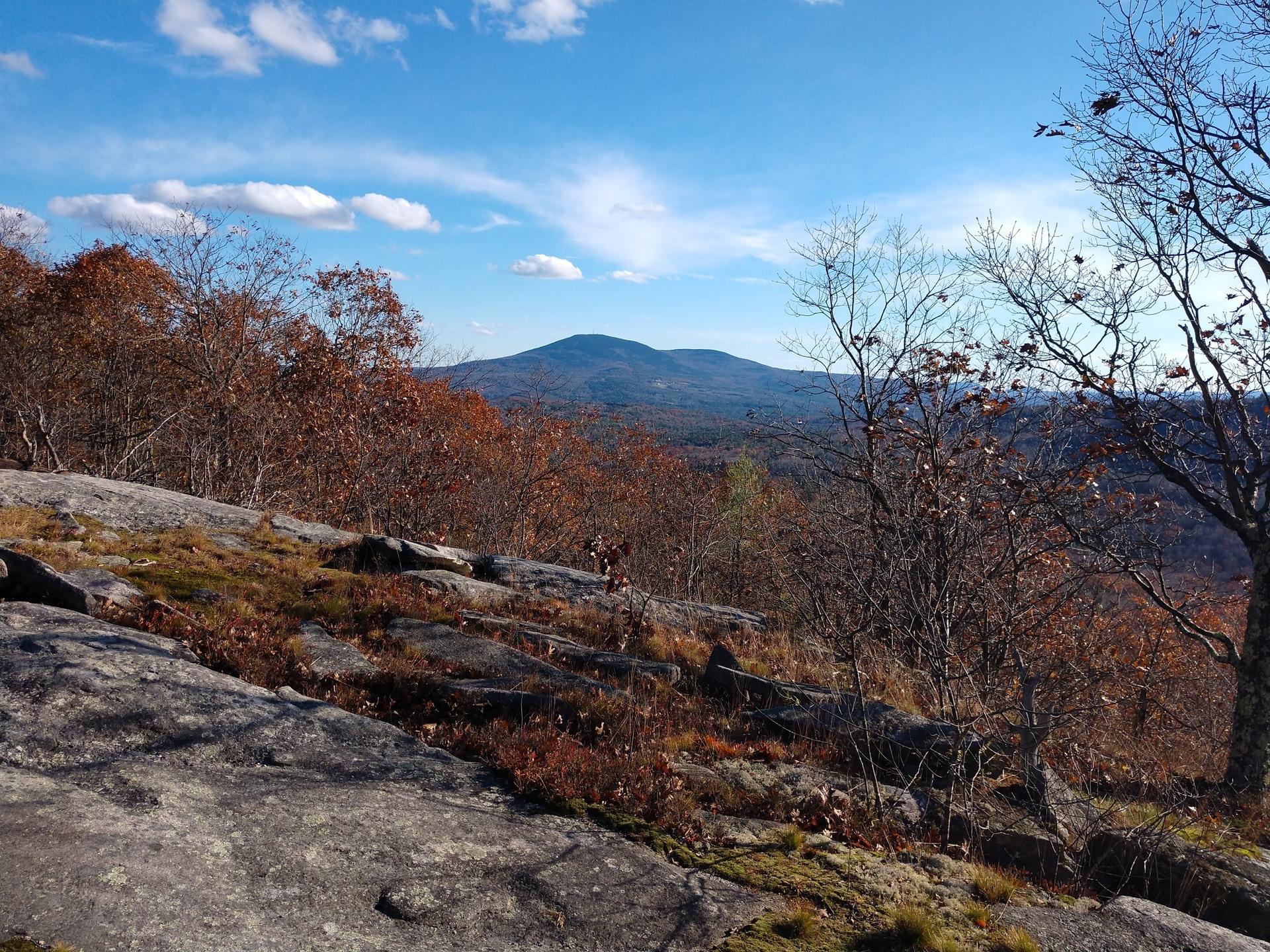 Sunny view of a rocky hillside, autumn trees, and a distant mountain under blue sky.