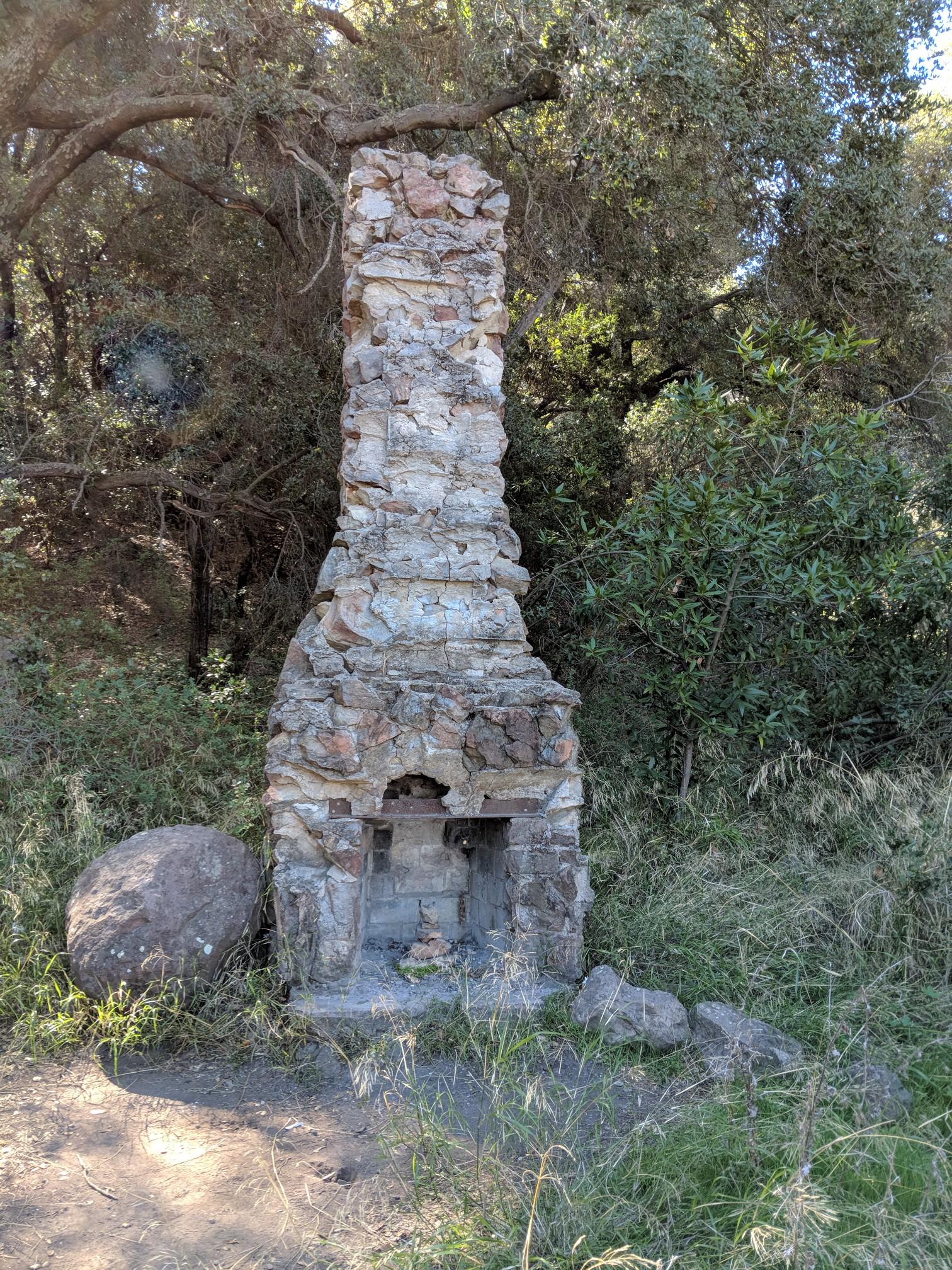 An old, tall stone chimney structure stands amidst green trees and dry brush.