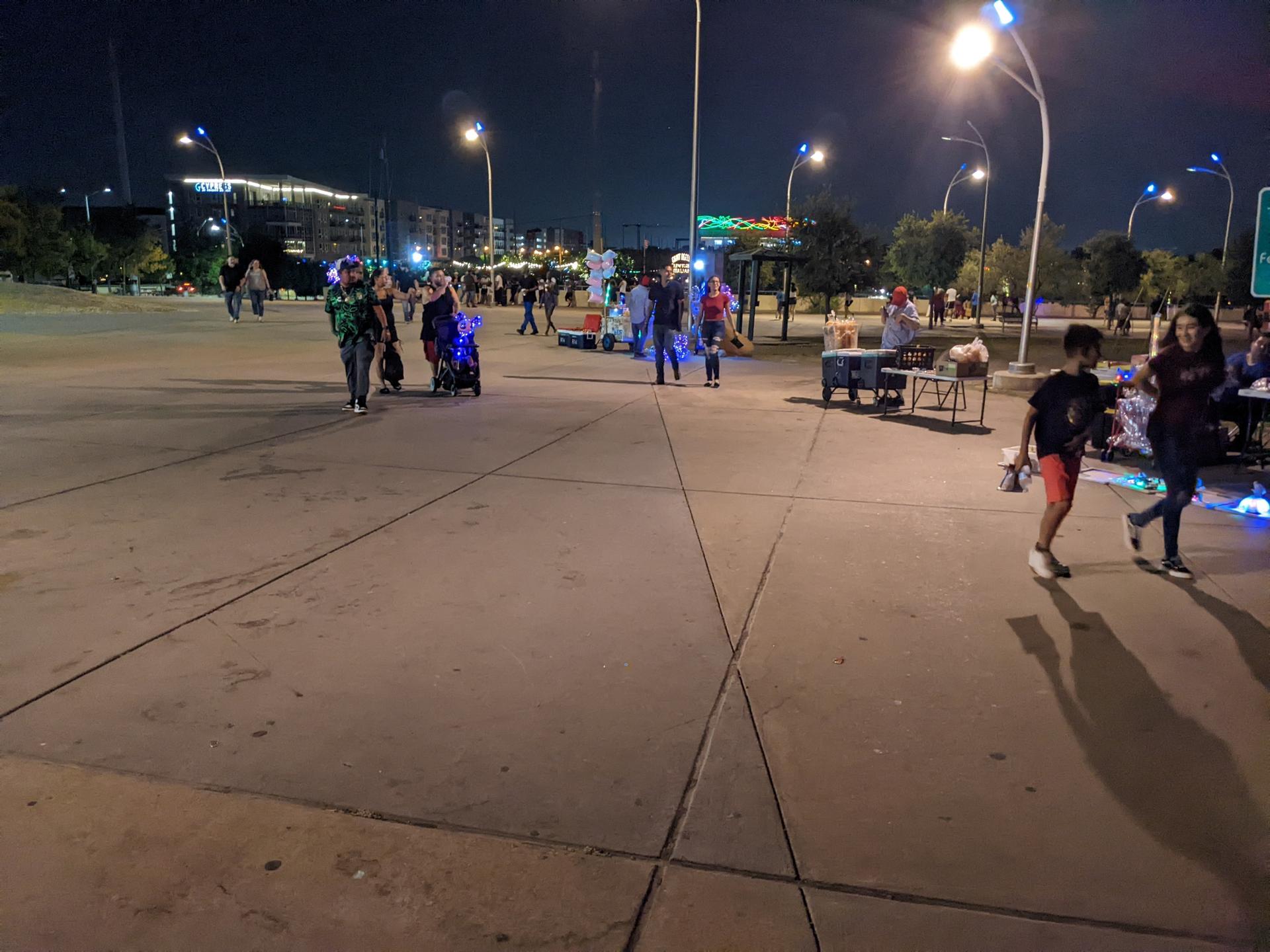 People in costumes walk on pavement at night under bright streetlights.