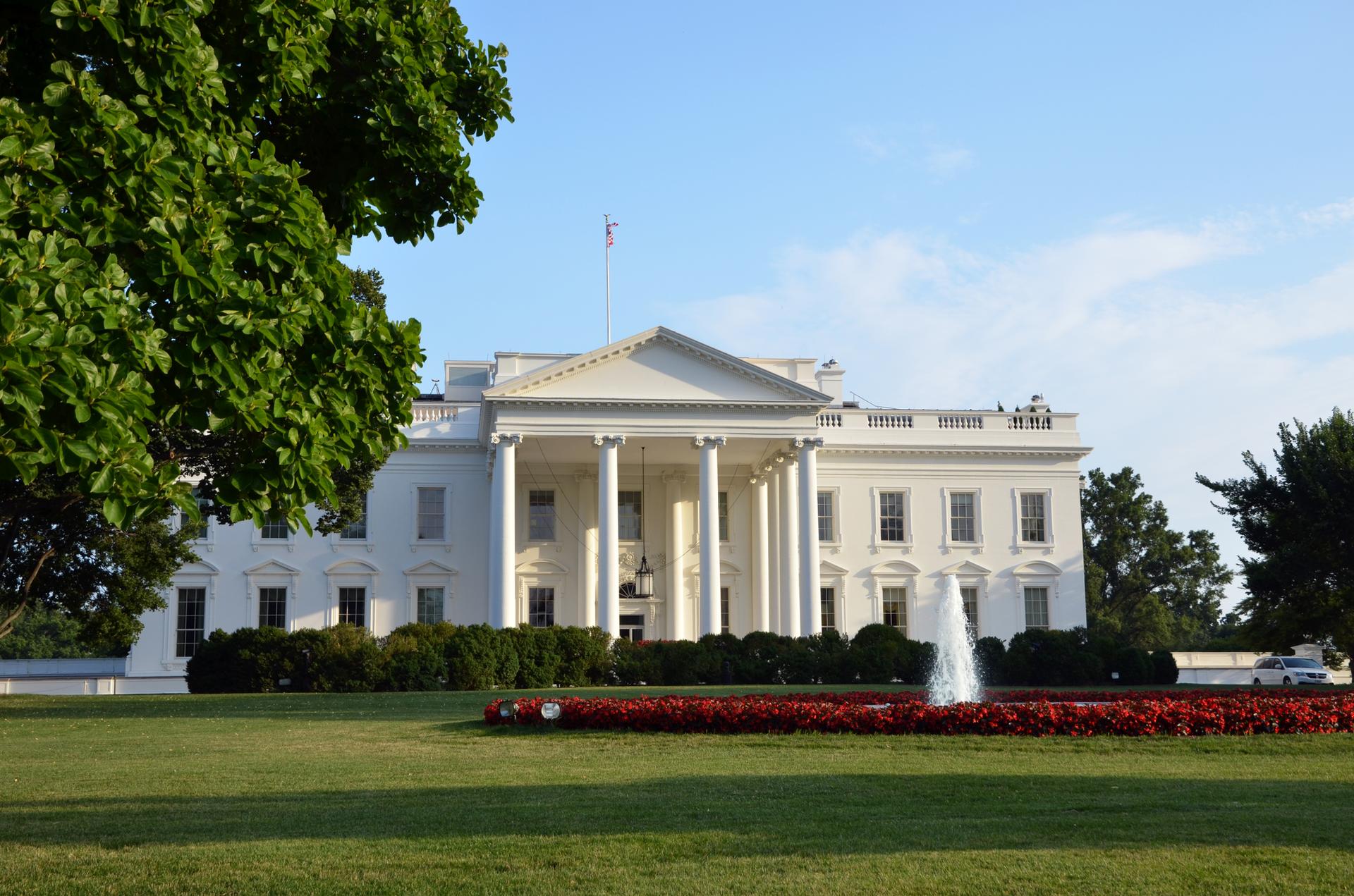 Grand white building with columns, fountain, and green lawn under clear sky.