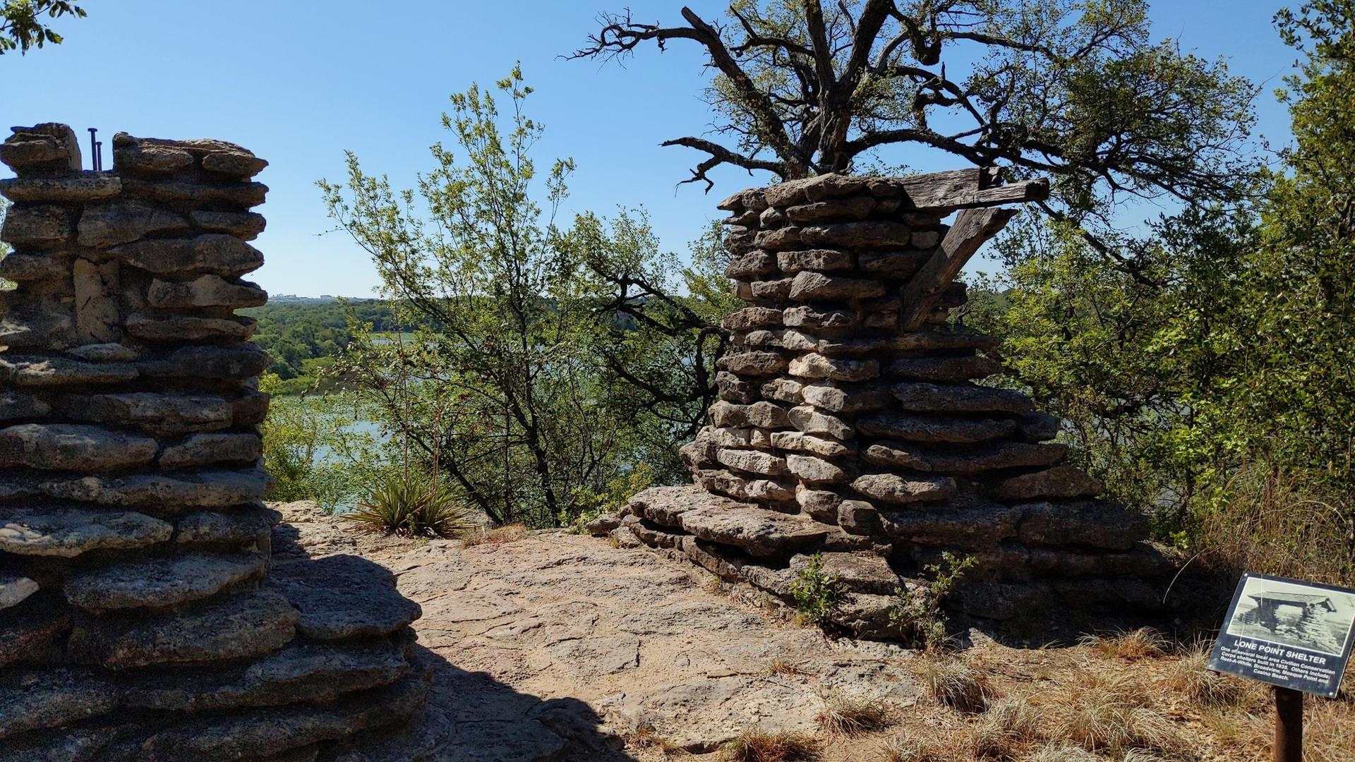 Two stacked stone structures overlook a sunny landscape with trees and water.