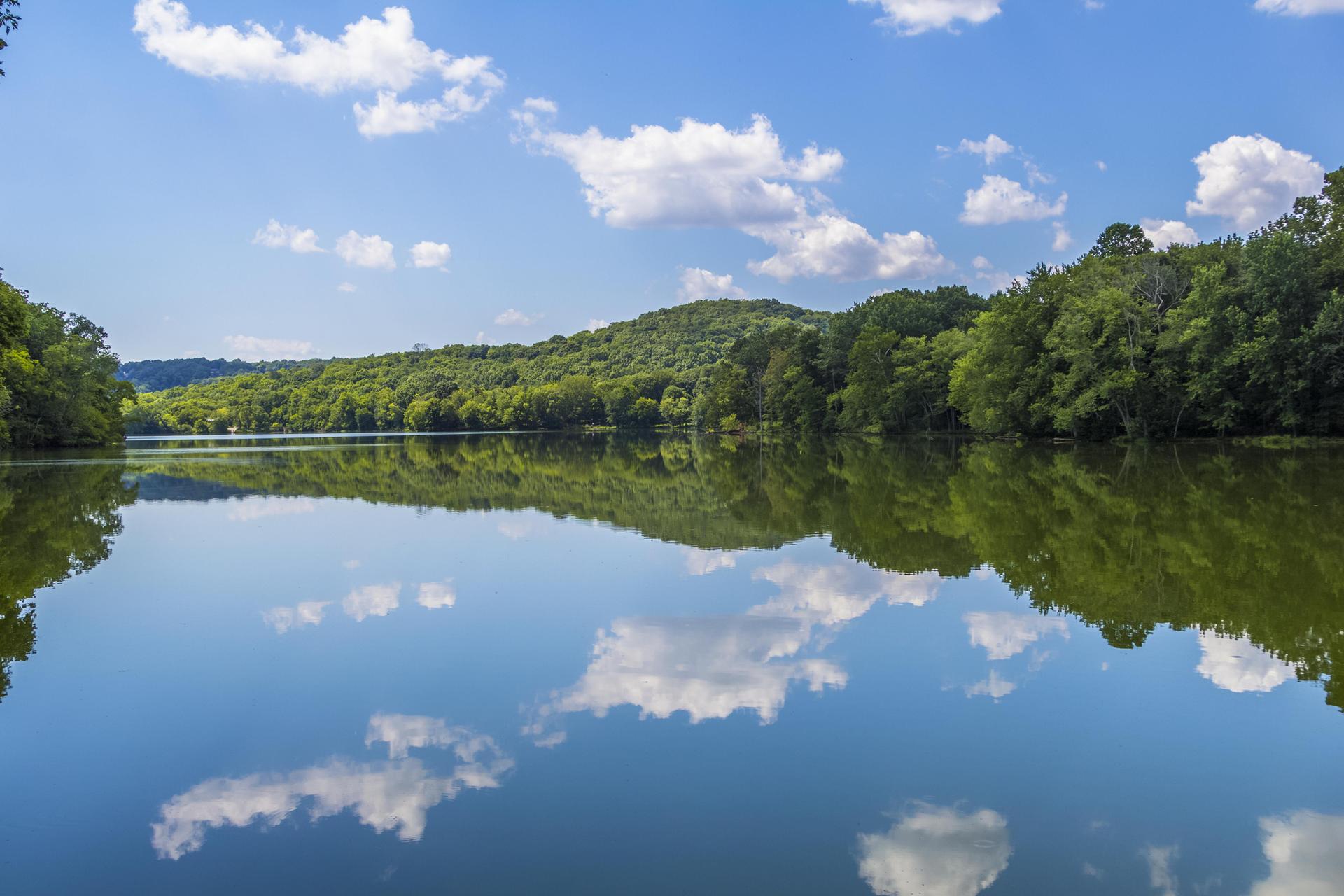 Wide lake reflecting blue sky and white clouds, surrounded by green forested hills.
