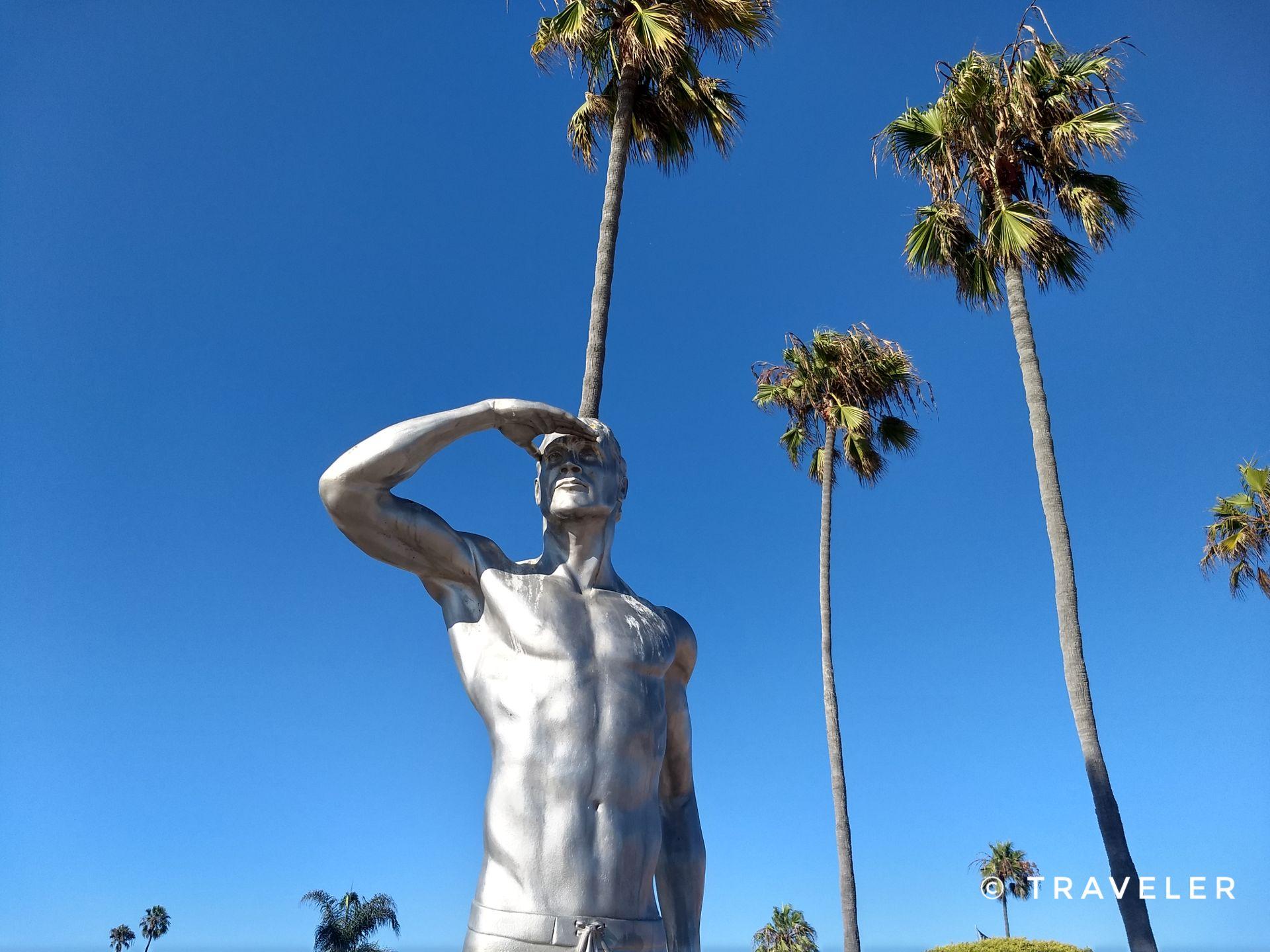 Silver statue of a man saluting, framed by tall palm trees.