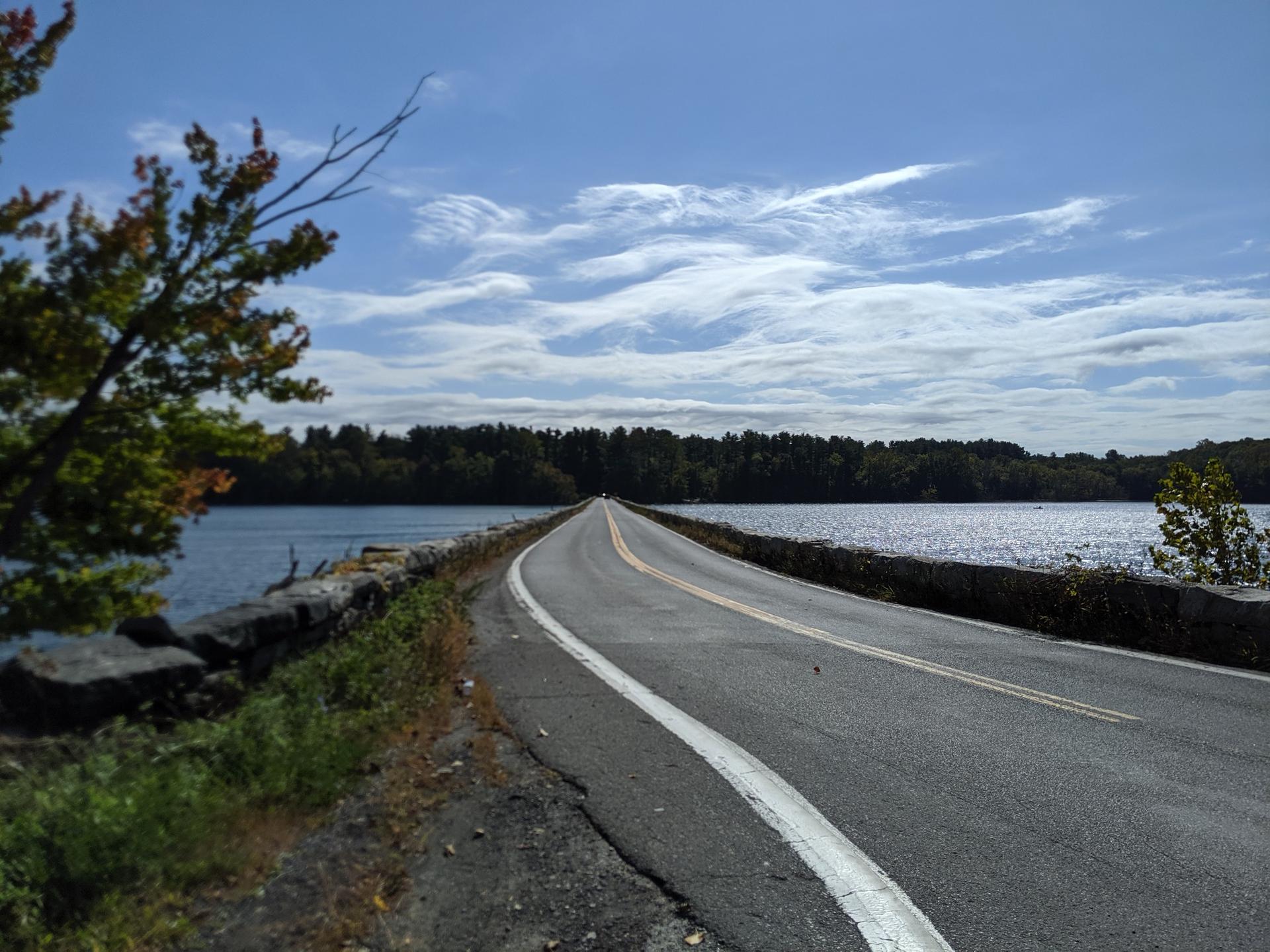 Road extends over a lake with forested shores under a cloudy sky.