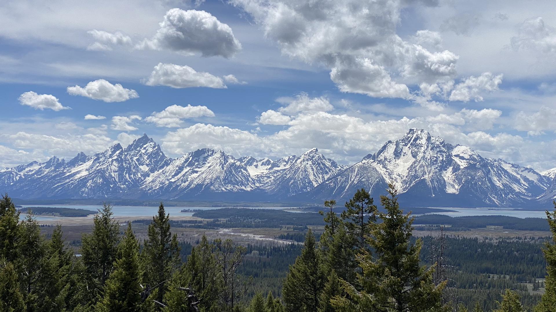 Snow-capped mountains behind a lake and forest under a partly cloudy sky.