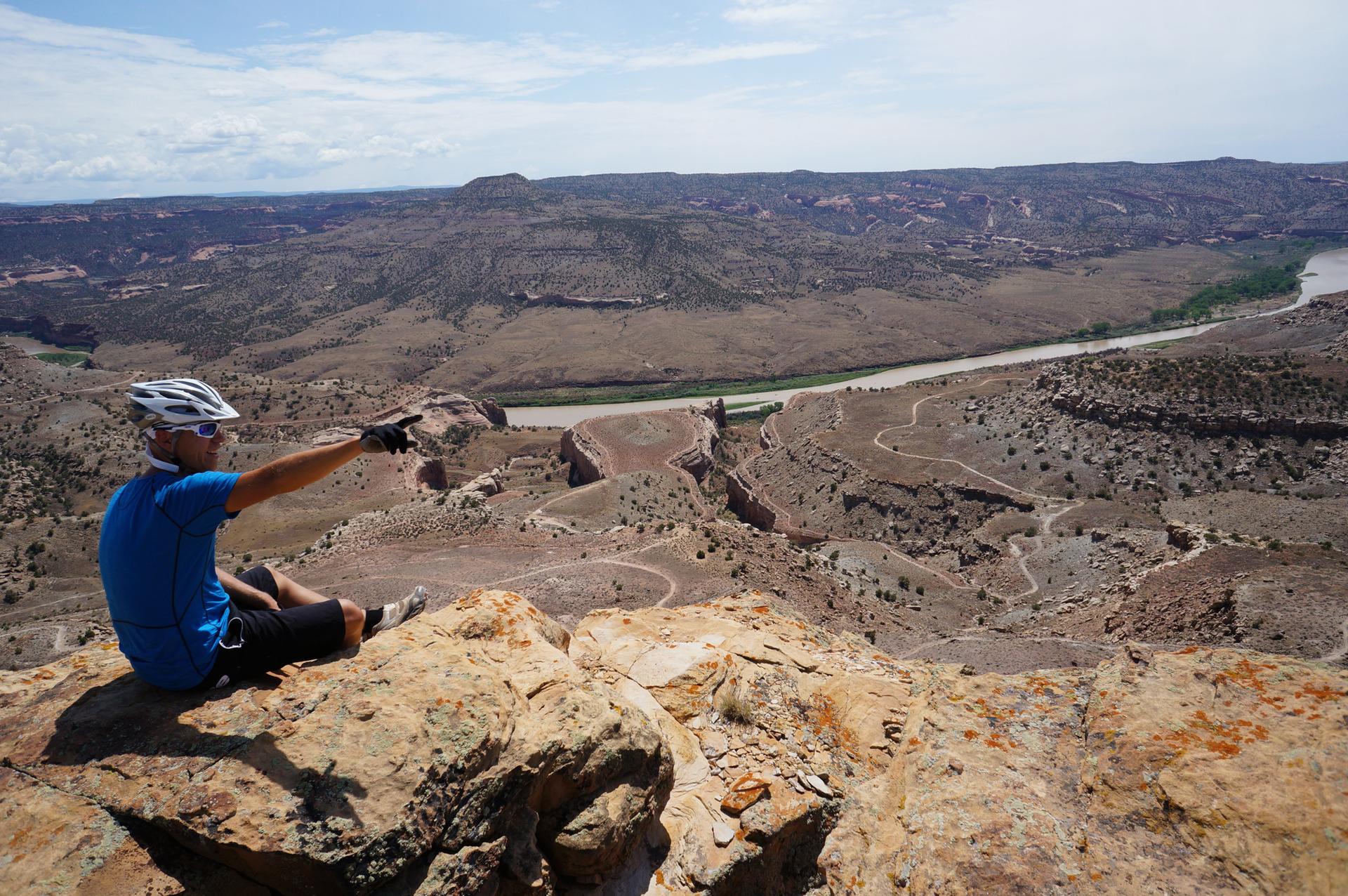 A man sits on a rock, pointing at a vast desert canyon view.