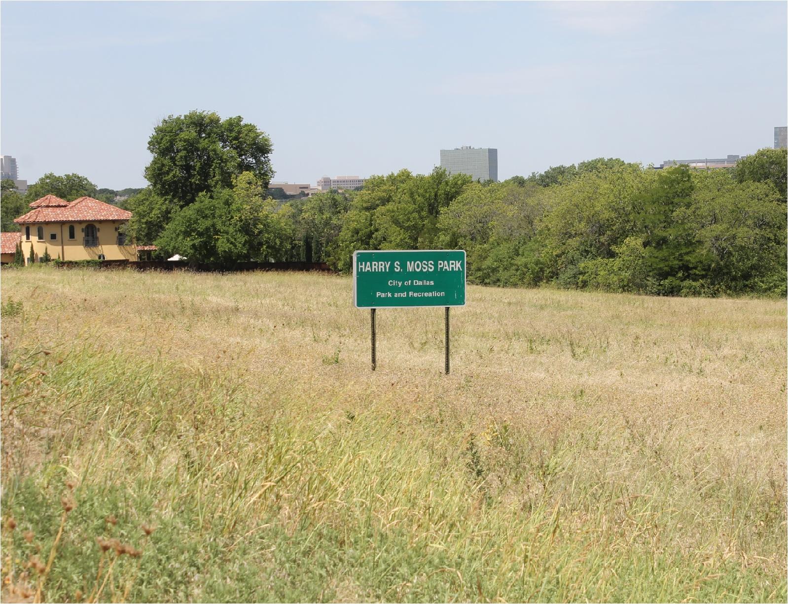 A green sign in a field of tall grass, with trees and buildings.
