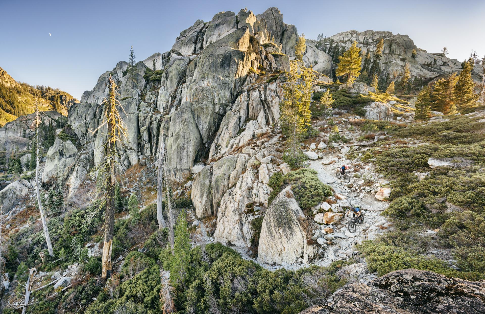 Two mountain bikers on a rocky trail winding through a steep, sunlit landscape.